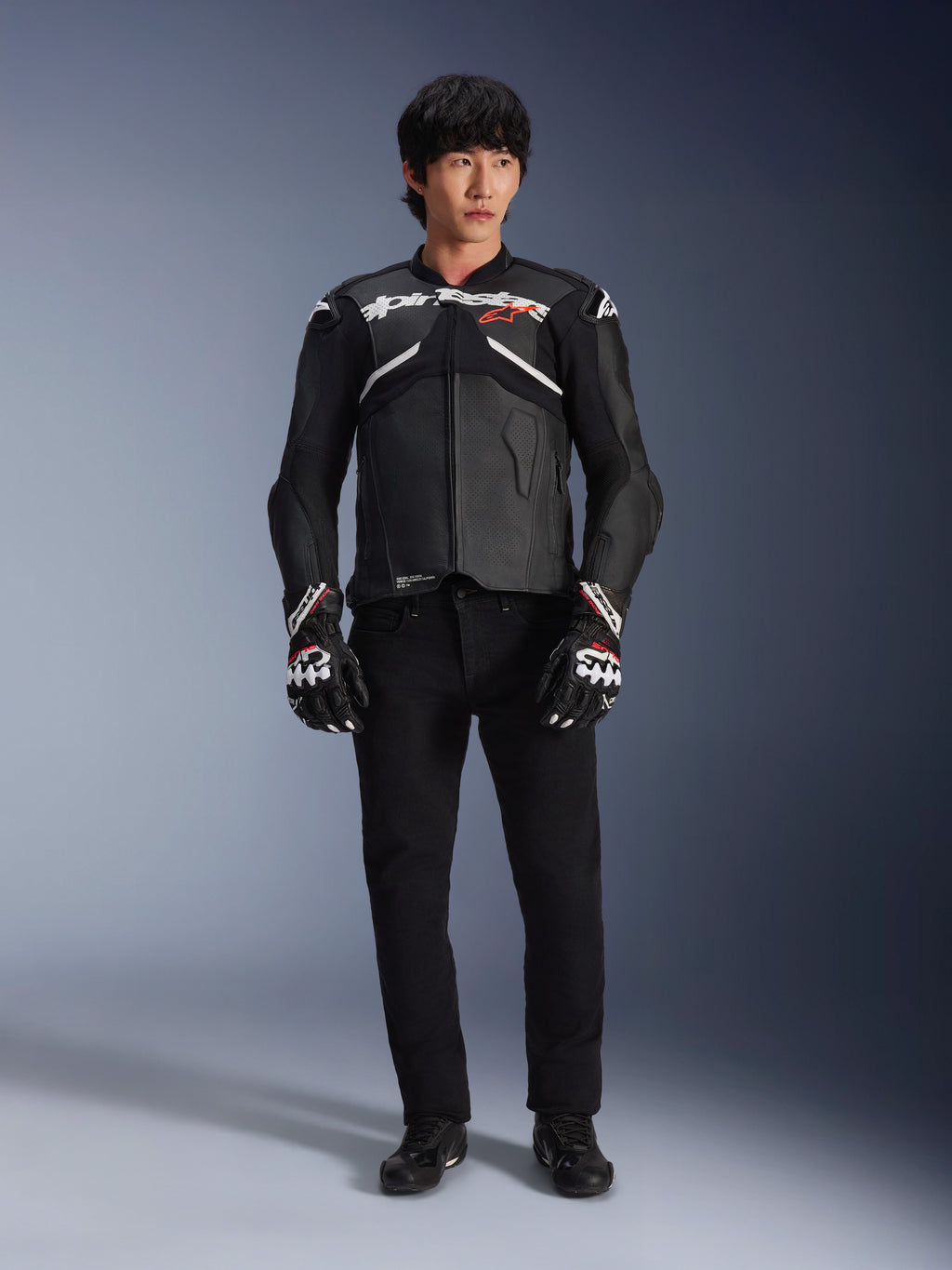 Model draagt de Alpinestars Atem V5 leren jas in zwart en wit, voorzien van witte Alpinestars-branding en een rood ster-logo op de borst, gecombineerd met een zwarte broek en zwart-witte racehandschoenen, strakke performance motoruitrusting