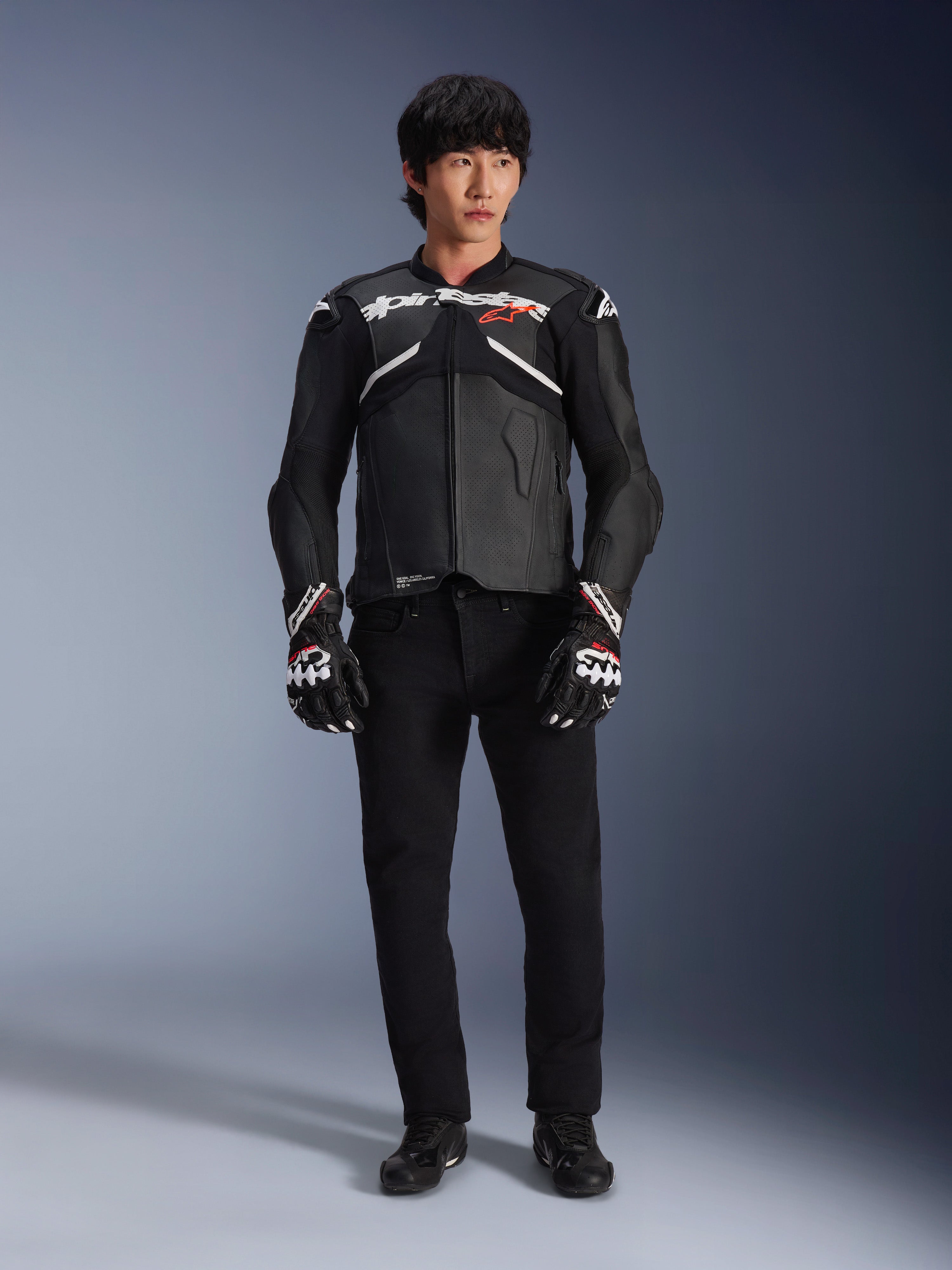 Model draagt de Alpinestars Atem V5 leren jas in zwart en wit, voorzien van witte Alpinestars-branding en een rood ster-logo op de borst, gecombineerd met een zwarte broek en zwart-witte racehandschoenen, strakke performance motoruitrusting