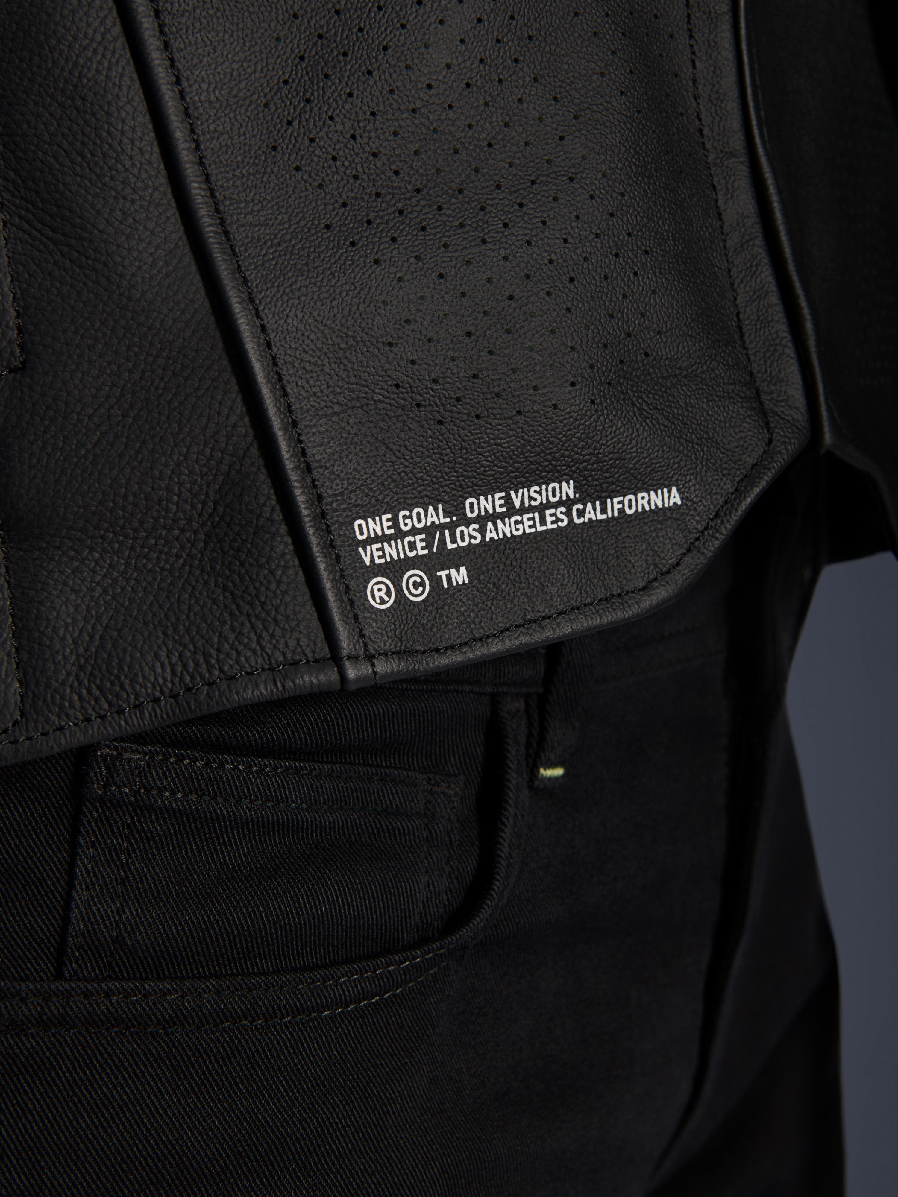 Close-up van de Alpinestars Atem V5 leren jas in het zwart, met het onderste rugpaneel voorzien van 'ONE GOAL. ONE VISION. VENICE / LOS ANGELES CALIFORNIA' branding en een geperforeerde leerstructuur.