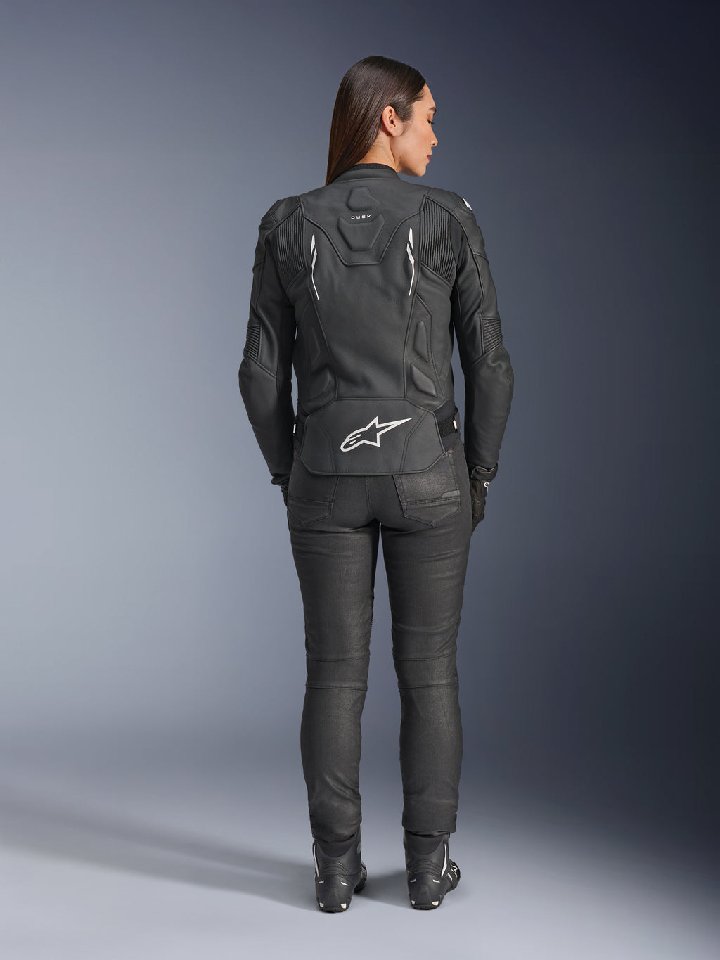 Alpinestars Stella Dusk leren jas voor dames, zwart, achteraanzicht met wit Alpinestars-logo op de onderrug en 'DUSK'-branding op de bovenrug, gedragen met zwarte motorbroek en laarzen, toont de aansluitende damespasvorm en beschermende leren constructie.