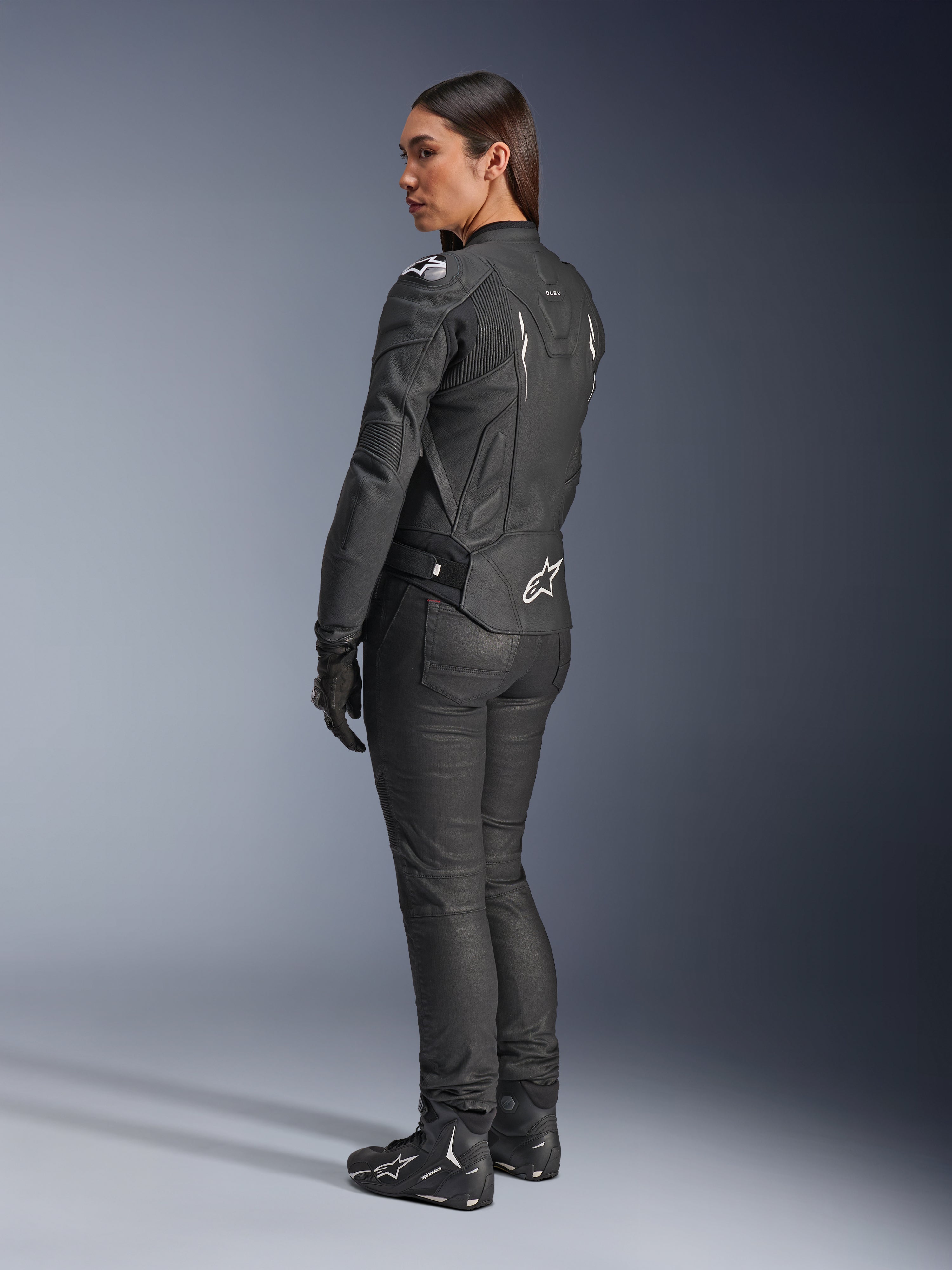 Alpinestars Stella Dusk leren jas, dames motorjas, zwart en wit, achteraanzicht met wit Alpinestars-logo op de onderrug en schouders, gedragen met zwarte motorbroek en laarzen, sportieve ergonomische pasvorm voor rijden op de weg