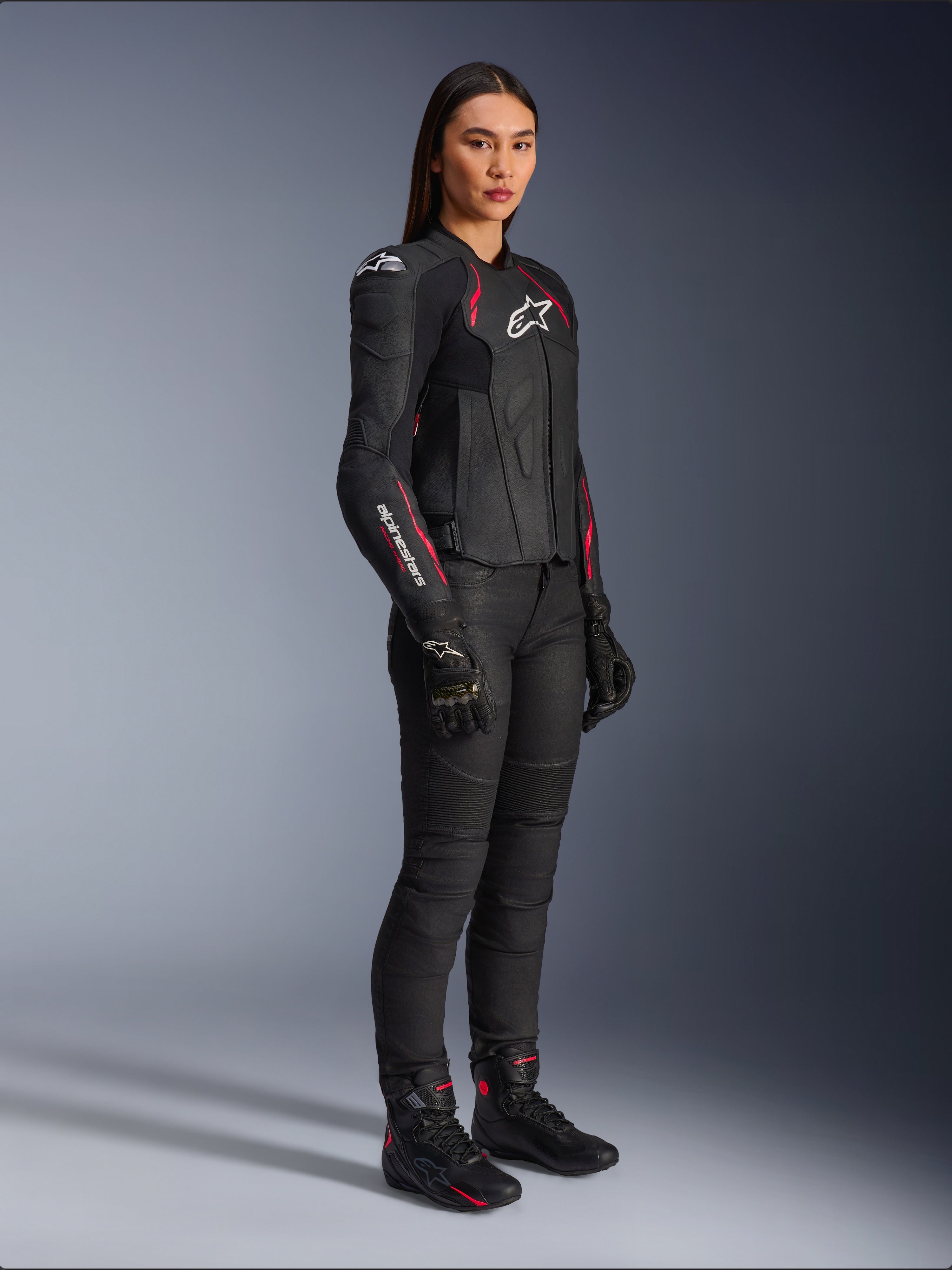 Alpinestars Stella Dusk leren damesjas, zwart met Diva roze en wit, gedragen door een vrouwelijk model met zwarte motorbroek en laarzen, voorzien van een strakke leren constructie met roze details en witte logo's
