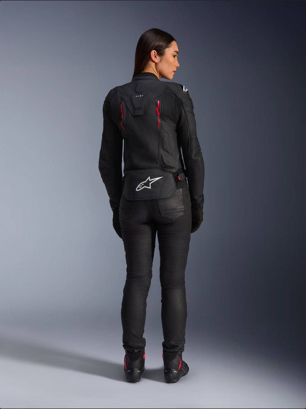 Alpinestars Stella Dusk leren motorjas, achteraanzicht, zwart leer met Diva Pink en witte accenten, voorzien van een wit Alpinestars-logo op de onderrug en roze ventilatiedetails, gedragen met een zwarte motorbroek en laarzen, hoogwaardige sportieve motoruitrusting voor dames