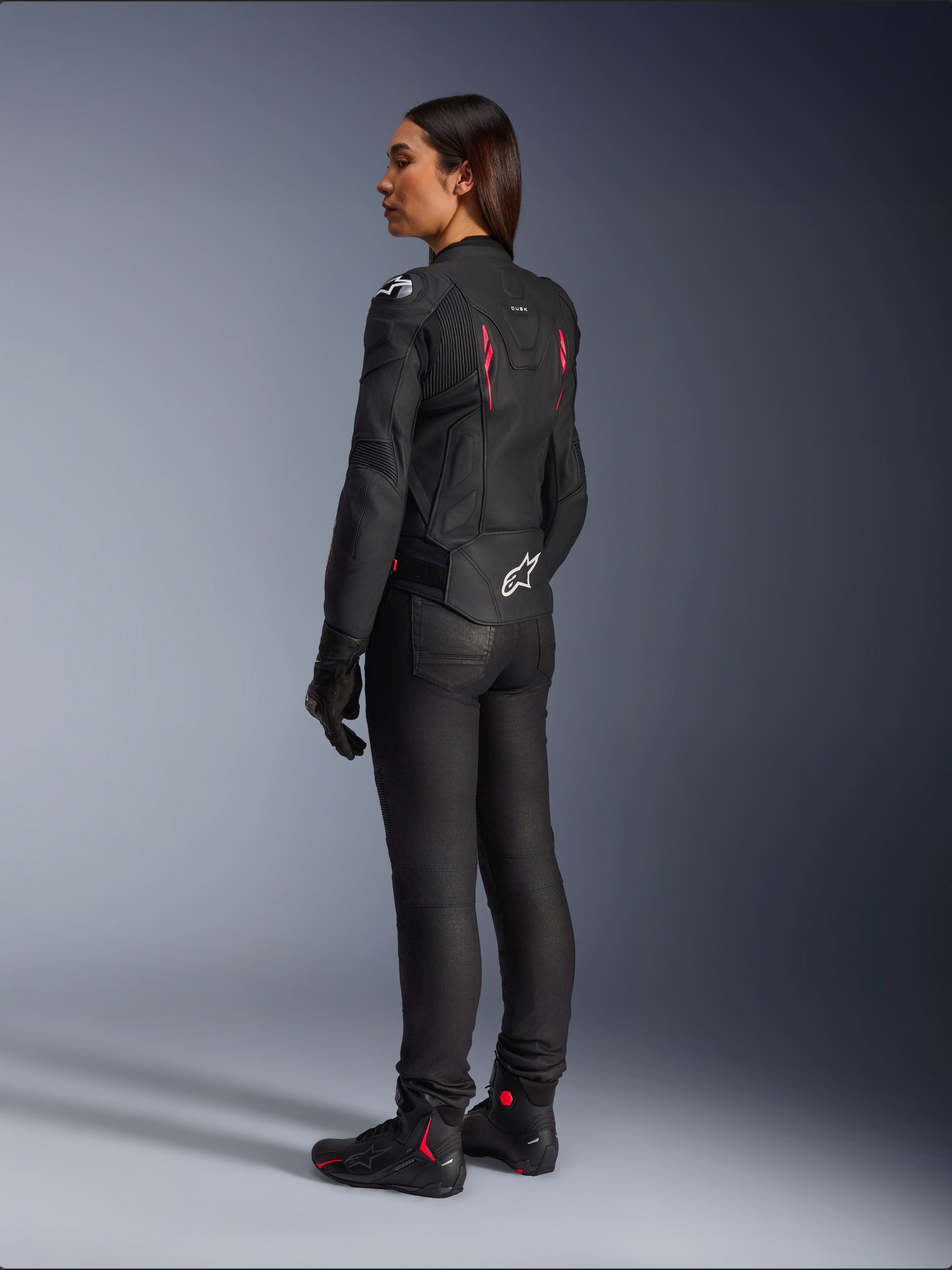 Alpinestars Stella Dusk dames leren jas, Black Diva Pink White, driekwart achteraanzicht op een rijder, gedragen met een zwarte motorbroek en zwarte laarzen, voorzien van een strakke leren constructie met roze accenten en een wit Alpinestars-logo op de onderrug.