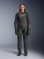 Alpinestars Stella Dusk leren jas, motorjas voor dames, Black Diva Pink White met wit logo op de borst en roze accenten op de schouders en mouwen, gecombineerd met zwarte motorbroek, handschoenen en laarzen, persoon draagt volledige motoruitrusting