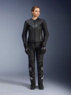 Alpinestars Stella GP Plus V4 leren motorjas voor dames in het zwart, gedragen door een vrouwelijke rijder met bijpassende leren racebroek, handschoenen en laarzen, voorzien van een getailleerde pasvorm, centrale rits en subtiel logo op de borst, afgestemd op sport- en wegmotorrijden.