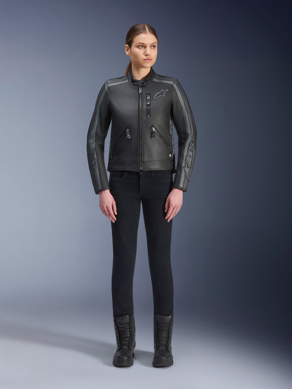 Alpinestars Stella Dyno Leren Jas, dames motorjas, zwart tar grijs donkergrijs, studio-opname van het volledige lichaam van een model dat de jas draagt met een zwarte slim-fit broek en zwarte laarzen, vervaardigd uit runderleer met grijze accenten en subtiele Alpinestars-branding op de borst en mouwen.