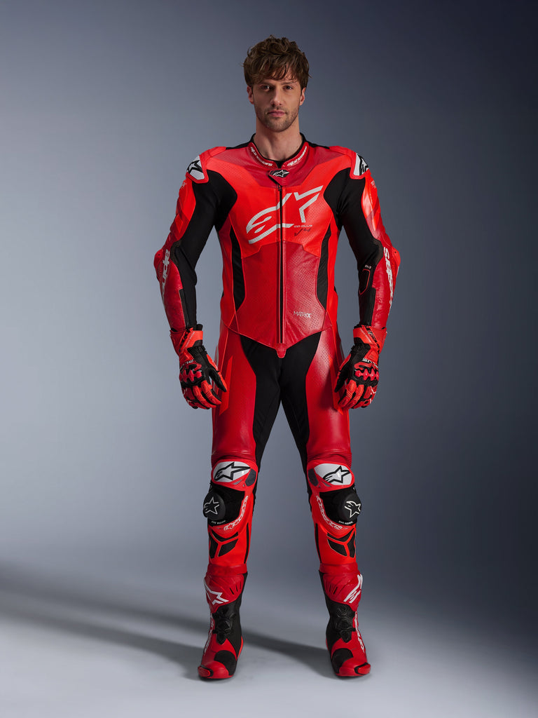 Model in Alpinestars GP Plus V4 Sprint eendelig leren pak, Red Fluo Mid Red en White, met zwarte en witte accenten, gecombineerd met bijpassende racehandschoenen en laarzen, sportief ontwerp met groot borstlogo en geperforeerde leren panelen voor ventilatie.