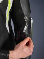 GP Tech V5 One Piece Leather Suit, detail view of the Tech-Air connection port on the sleeve, hand demonstrating electronic integration, black leather with yellow fluo and white perforated panels, professional racing gear