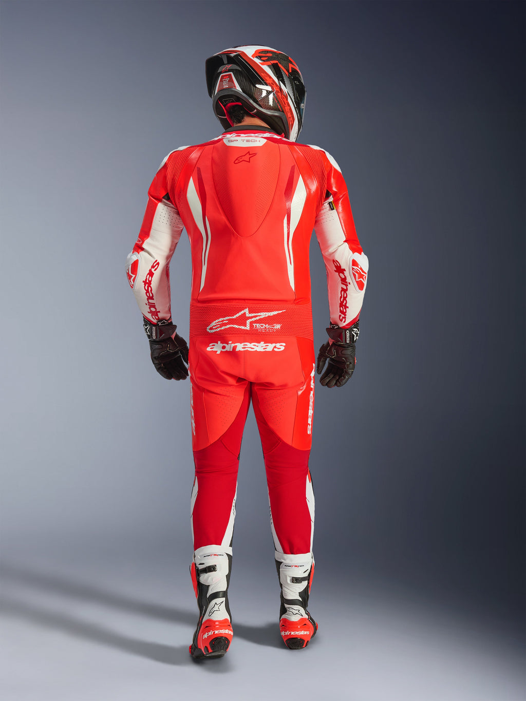 Achteraanzicht van een rijder in het Alpinestars GP Tech V5 eendelig leren pak in Red Fluo, Bright Red en White. Hoogwaardig leren pak met een aerodynamische speedhump, witte accenten en Alpinestars-branding, gecombineerd met een bijpassende helm, zwarte handschoenen en wit-rode racelaarzen.
