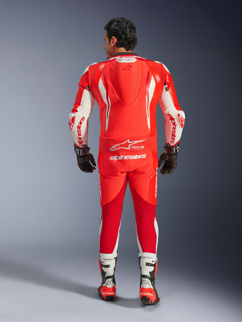 GP Tech V5 Eendelig Leren Pak, motorracepak, Red Fluo Bright Red en White, achteraanzicht met aerodynamische bult en witte Alpinestars-branding, gedragen met zwart-witte racehandschoenen en laarzen.