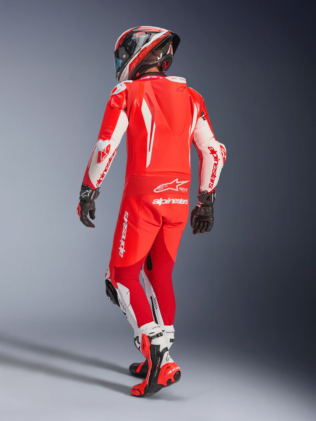 Alpinestars GP Tech V5 eendelig leren pak, Red Fluo Bright Red White, achteraanzicht van een motorrijder in volledige race-uitrusting, met een aerodynamische bult en witte panelen op de mouwen en pijpen, gecombineerd met een bijpassende helm en rode racelaarzen, professioneel ontwerp voor circuitprestaties.