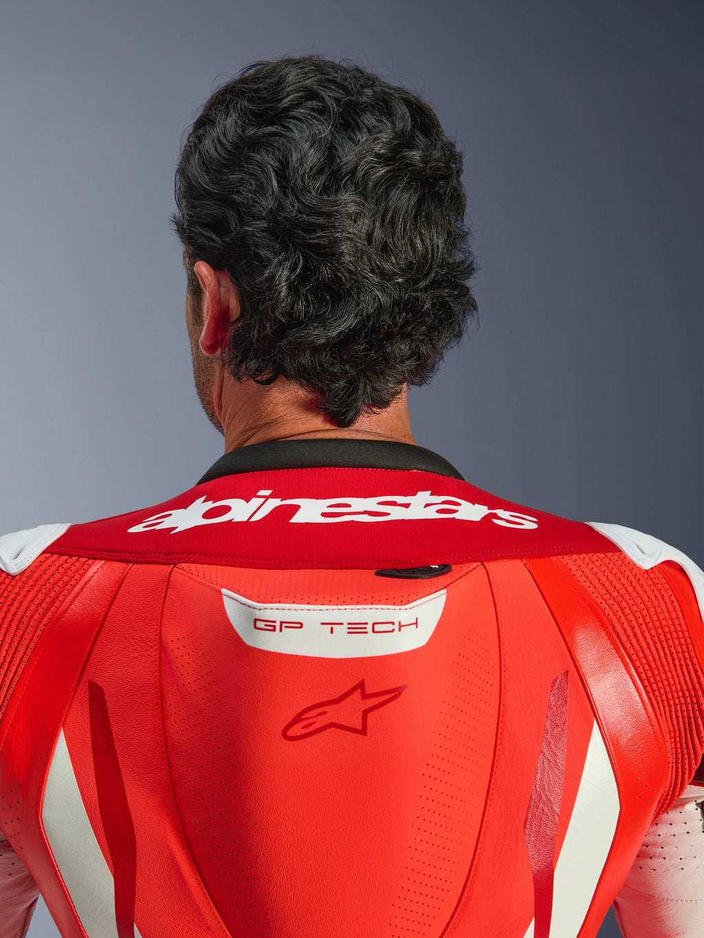 Close-up van de bovenrug en nek van het GP Tech V5 eendelig leren pak, Red Fluo en Bright Red, met wit Alpinestars-woordmerk op de kraag en GP TECH-logo op de aerodynamische bult, gedragen door een rijder.