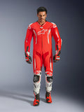 Alpinestars GP Tech V5 eendelig lederen motorpak, professioneel racepak in rood fluo en helderrood met witte accenten, gedragen door een rijder met bijpassende laarzen en handschoenen, voorzien van een aerodynamische snelheidsbult, hoogwaardige lederen constructie en geavanceerde bescherming voor circuitprestaties.