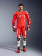 Alpinestars GP Tech V5 eendelig lederen motorpak, professioneel racepak in rood fluo en helderrood met witte accenten, gedragen door een rijder met bijpassende laarzen en handschoenen, voorzien van een aerodynamische snelheidsbult, hoogwaardige lederen constructie en geavanceerde bescherming voor circuitprestaties.