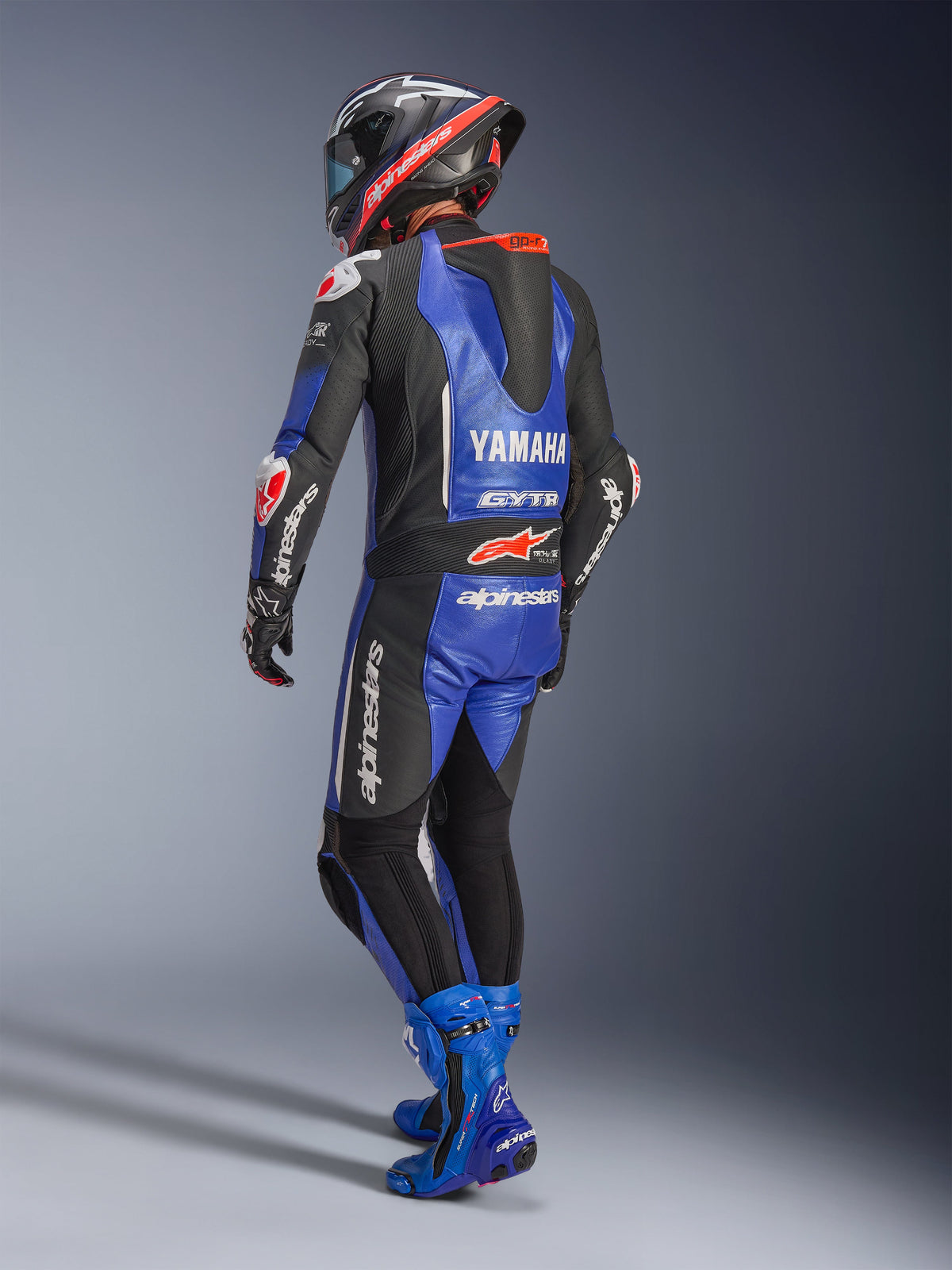 Alpinestars Yamaha GP-R7 eendelig leren pak, metallic blauw, zwart en wit, achteraanzicht met aerodynamische racebult en Yamaha-branding, gedragen met bijpassende blauwe racelaarzen en helm, professioneel leren performance-pak voor op het circuit