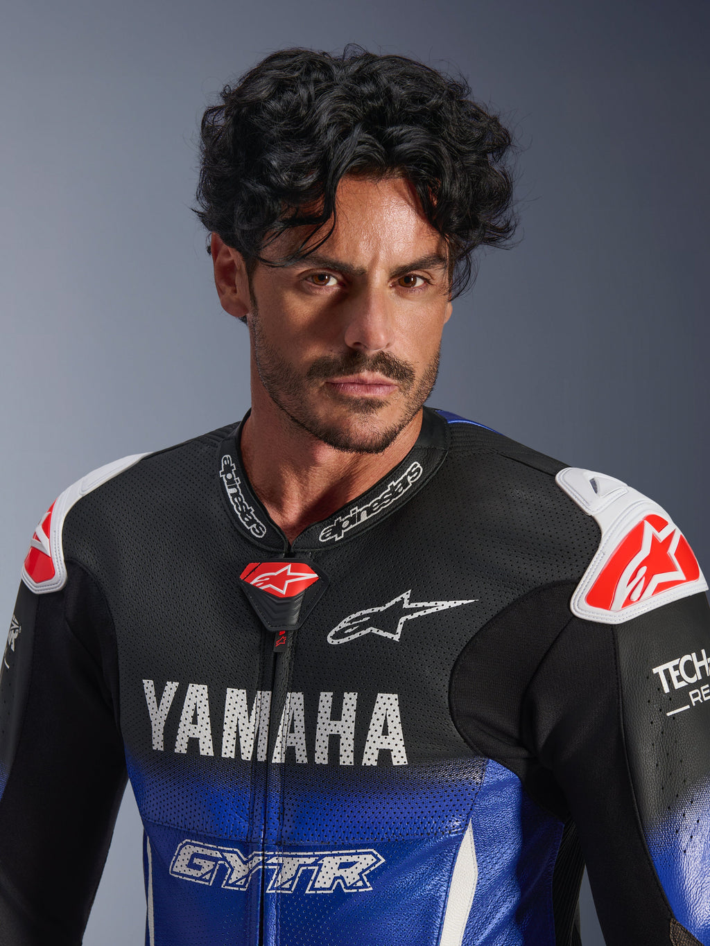Close-up portret van een man in het Alpinestars Yamaha GP-R7 eendelig leren pak, metallic blauw en zwart met witte YAMAHA- en GYTR-branding, geperforeerd leer, witte schouder sliders met rode ster-logo's, Tech-Air Ready, professionele motorrace-uitrusting.