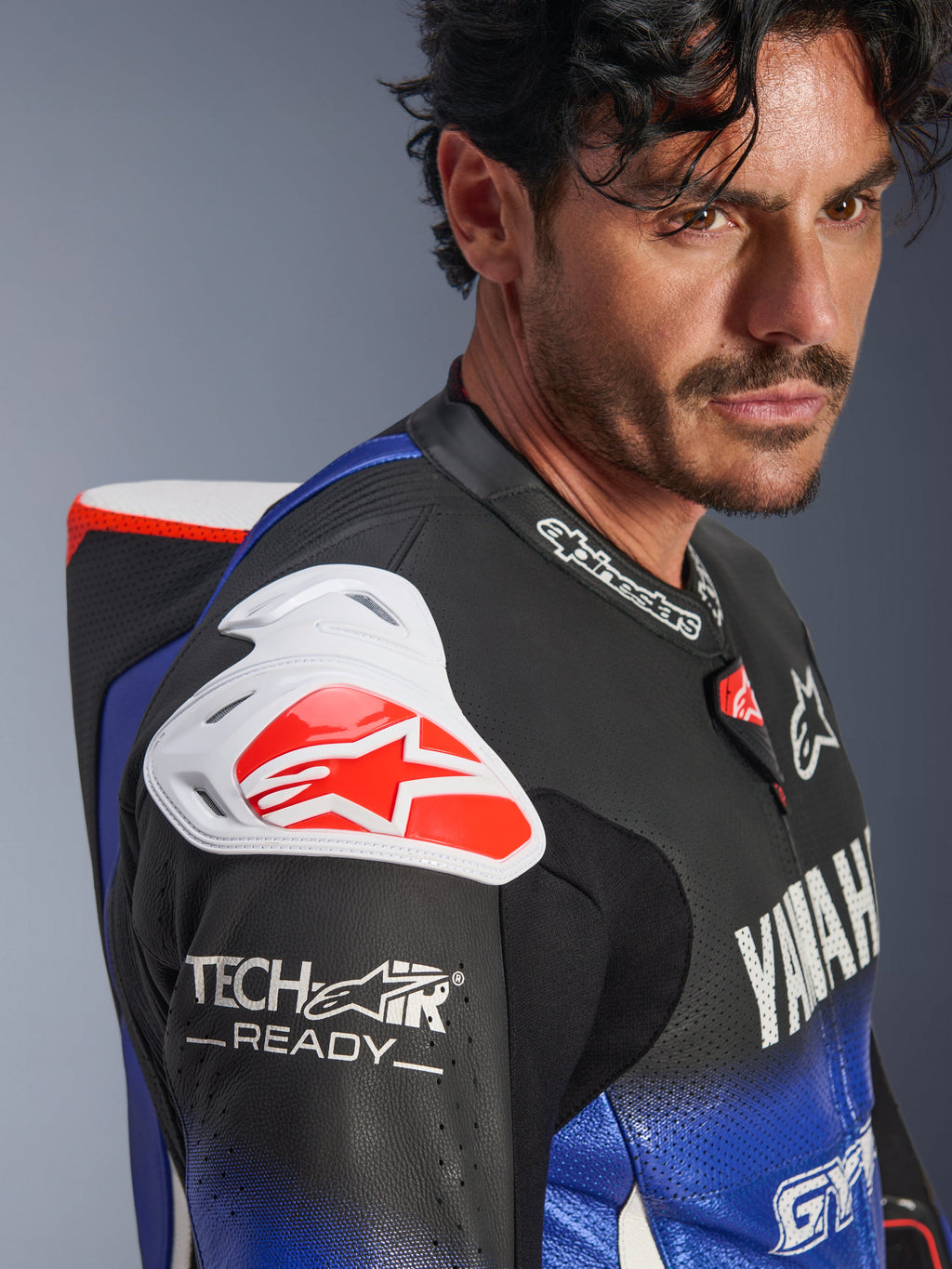 Yamaha GP-R7 Eendelig Leren Pak, metallic blauw, zwart, wit en rood, close-up van de schouder en borst, voorzien van witte DFS-schouderprotectie met een rood logo, aerodynamische bult en Tech-Air Ready-branding op de geperforeerde leren mouw.