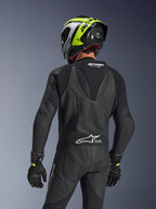 Alpinestars GP Force V2 Leather Suit 1-Piece, Black, rear view showing aerodynamic speed hump and perforated leather construction, worn with a black and neon yellow racing helmet and black gloves, featuring accordion stretch panels and white Tech-Air Ready branding.