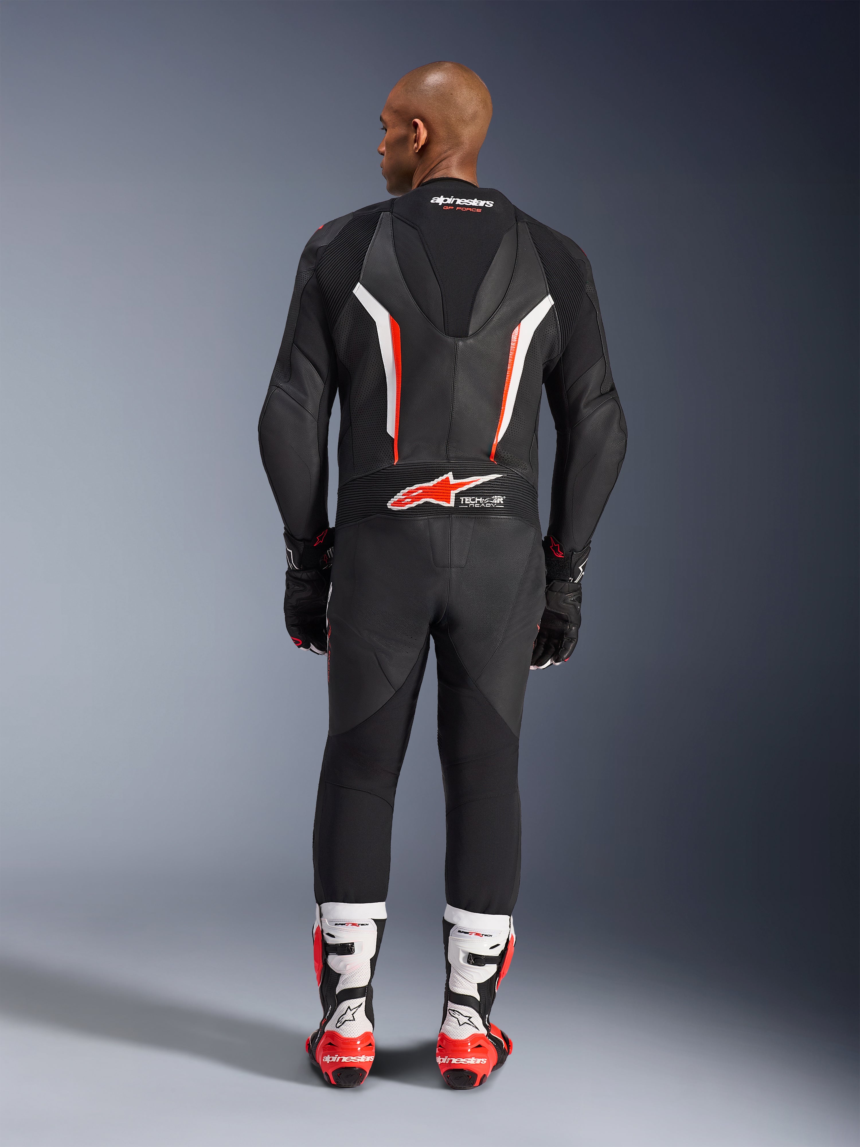 Achteraanzicht van een model in het Alpinestars GP Force V2 1-delig leren pak in Black White Red Fluo, met de aerodynamische speedhump en Tech-Air Ready-branding, gecombineerd met wit-rode professionele racelaarzen en zwarte handschoenen.