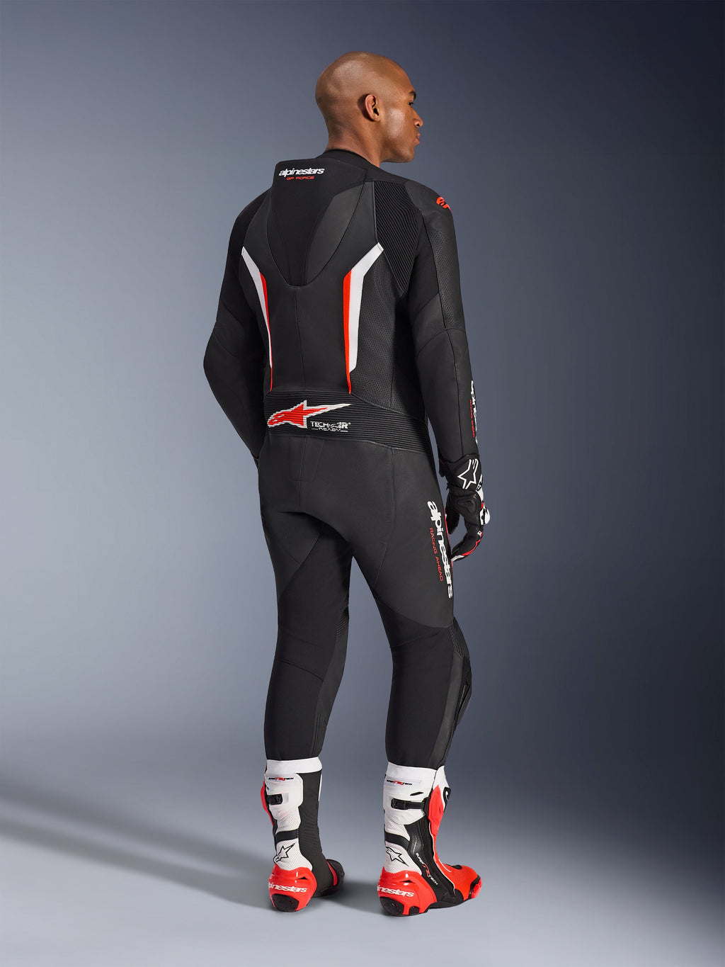 Motorrijder die het GP Force V2 Leren Pak 1-delig draagt, zwart met witte en rood fluo accenten, van achteren gezien met de aerodynamische speedhump en Alpinestars-branding, gecombineerd met bijpassende racehandschoenen en witte en rode laarzen, professionele motorrace-uitrusting