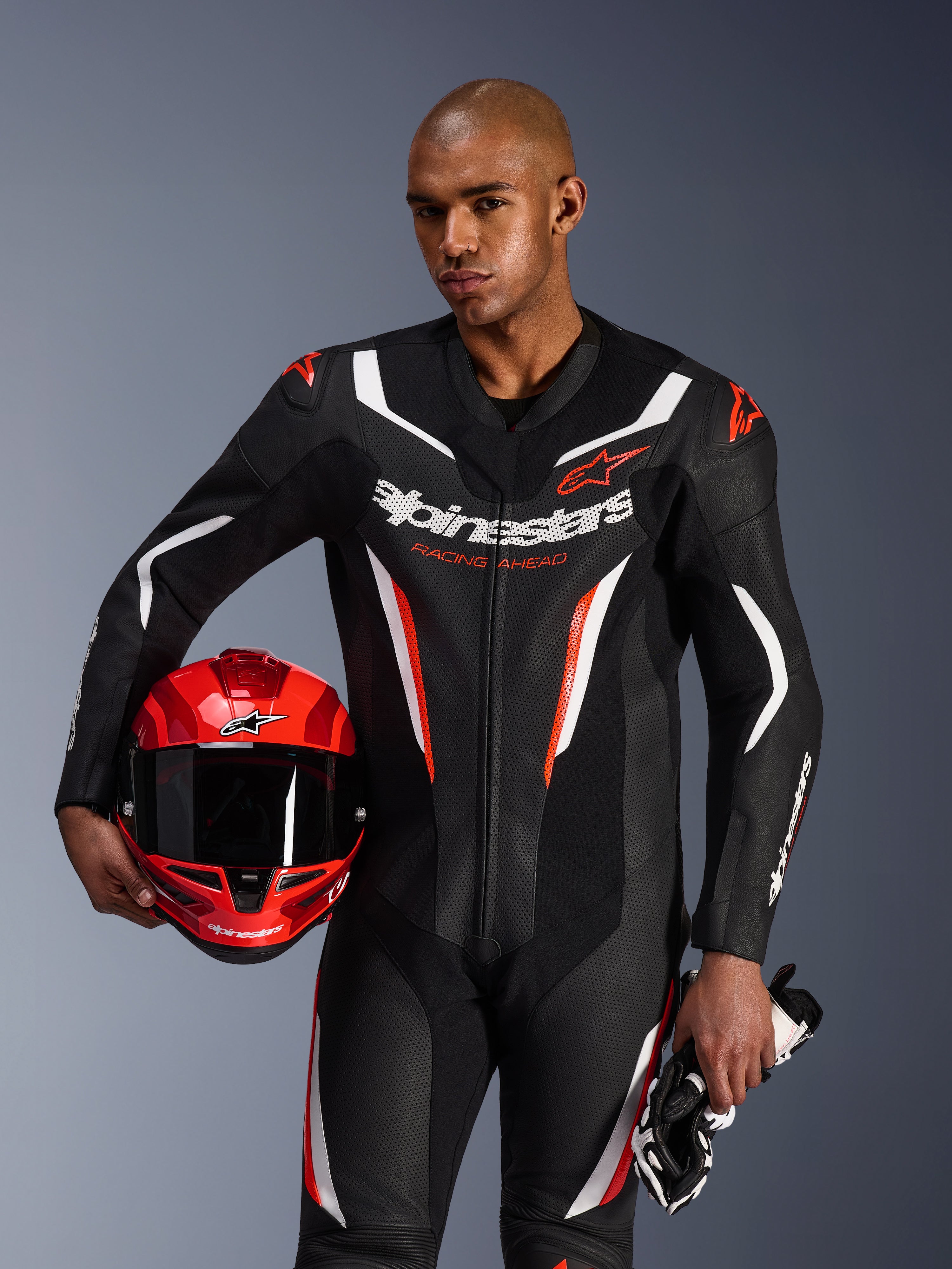 Alpinestars GP Force V2 Leren Pak, 1-delig motorpak, zwart, wit en fluorood, gedragen door een model dat een rode helm en handschoenen vasthoudt, strak race-ontwerp met geperforeerd leer en opvallende branding
