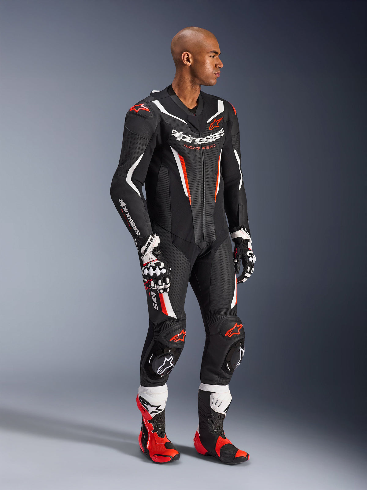 Model draagt het Alpinestars GP Force V2 1-delig leren pak in Black White Red Fluo, hoogwaardige race-uitrusting met geperforeerde leren panelen en DFS Lite-schilden, gecombineerd met bijpassende rood-witte racelaarzen en handschoenen, professionele motorsport-uitstraling.