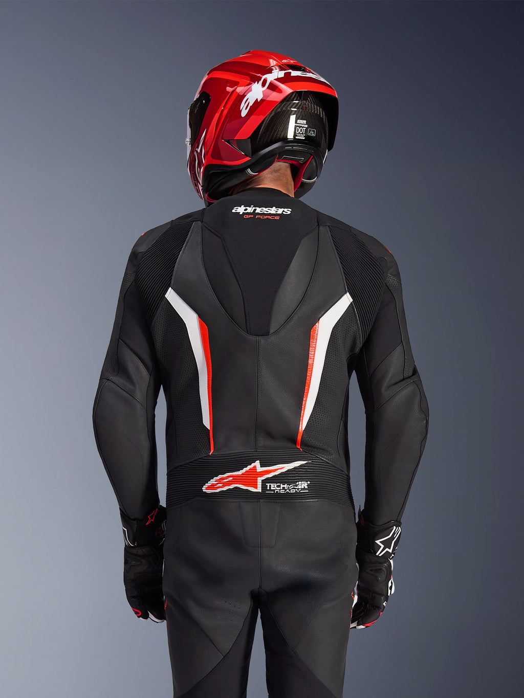 Alpinestars GP Force V2 Leather Suit 1-Piece, zwart wit en rood fluo, achteraanzicht van rijder met rode racehelm en handschoenen, voorzien van geperforeerde leren panelen, aerodynamische speedhump met Alpinestars-branding en Tech-Air Ready-logo op de onderrug, professionele motorrace-uitrusting.