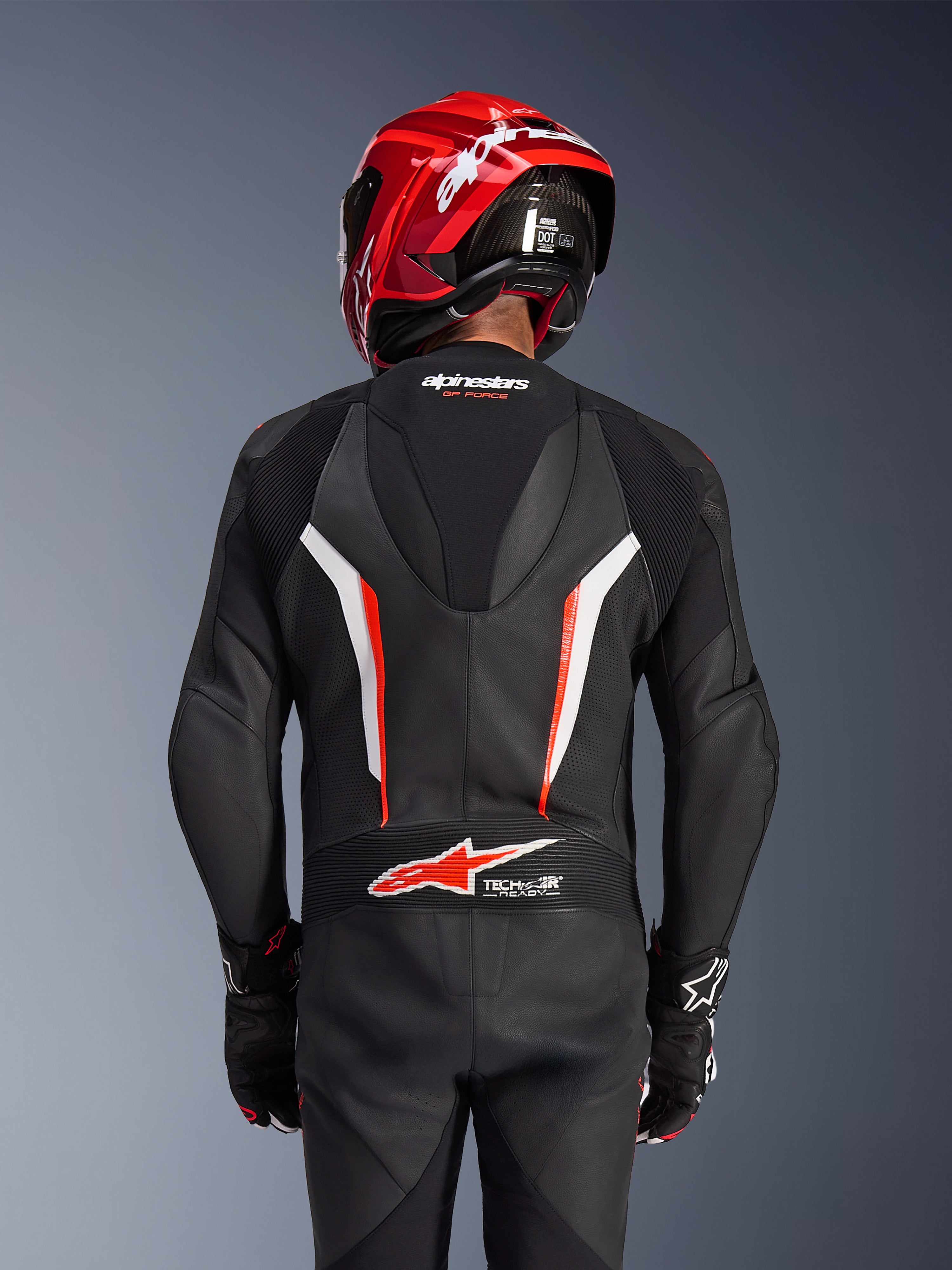 Alpinestars GP Force V2 Leather Suit 1-Piece, zwart wit en rood fluo, achteraanzicht van rijder met rode racehelm en handschoenen, voorzien van geperforeerde leren panelen, aerodynamische speedhump met Alpinestars-branding en Tech-Air Ready-logo op de onderrug, professionele motorrace-uitrusting.