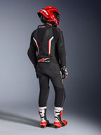 Motorrijder van achteren gezien in het Alpinestars GP Force V2 1-delig leren pak in zwart, wit en fluorood, met een aerodynamische speedhump en geperforeerde leren panelen, gecombineerd met een rode Alpinestars helm, racehandschoenen en rood-witte motorlaarzen, professionele circuit-raceuitrusting.