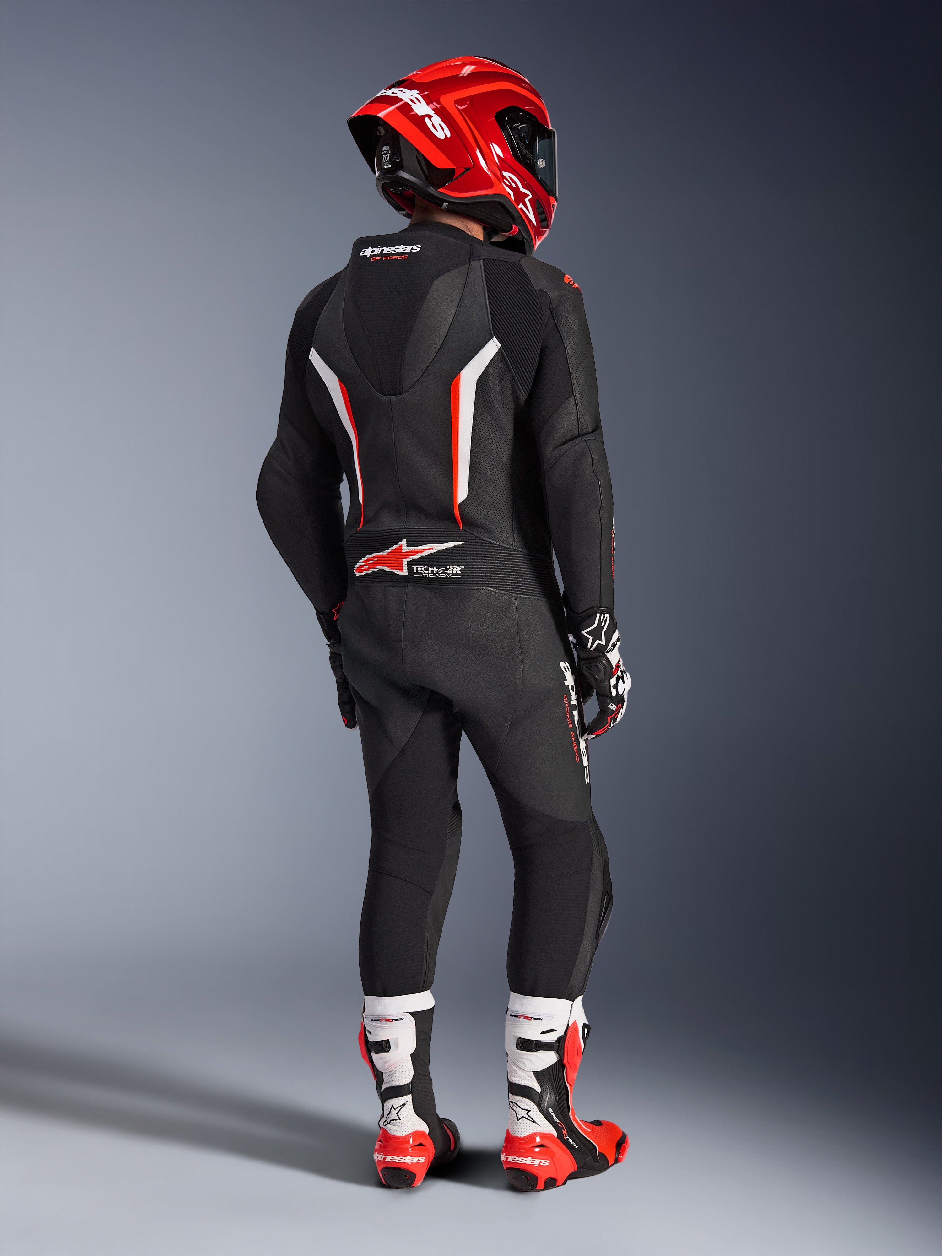 Motorrijder van achteren gezien in het Alpinestars GP Force V2 1-delig leren pak in zwart, wit en fluorood, met een aerodynamische speedhump en geperforeerde leren panelen, gecombineerd met een rode Alpinestars helm, racehandschoenen en rood-witte motorlaarzen, professionele circuit-raceuitrusting.