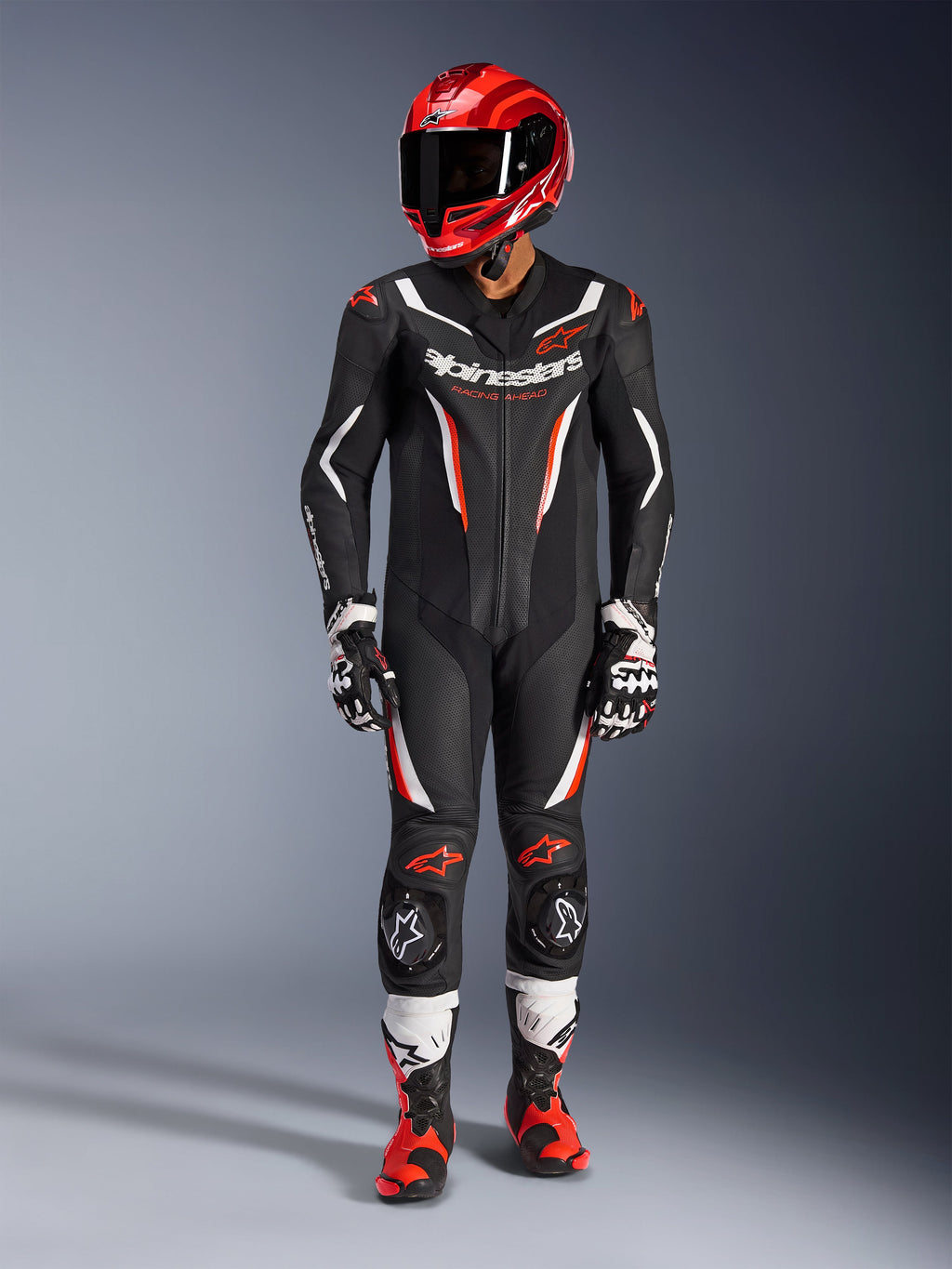Alpinestars GP Force V2 Leren Pak - 1-delig, Black White Red Fluo, gedragen met een rode racehelm, zwart-witte handschoenen en rood-zwarte laarzen, met geperforeerde leren panelen en Alpinestars-branding, professionele motorrace-uitrusting