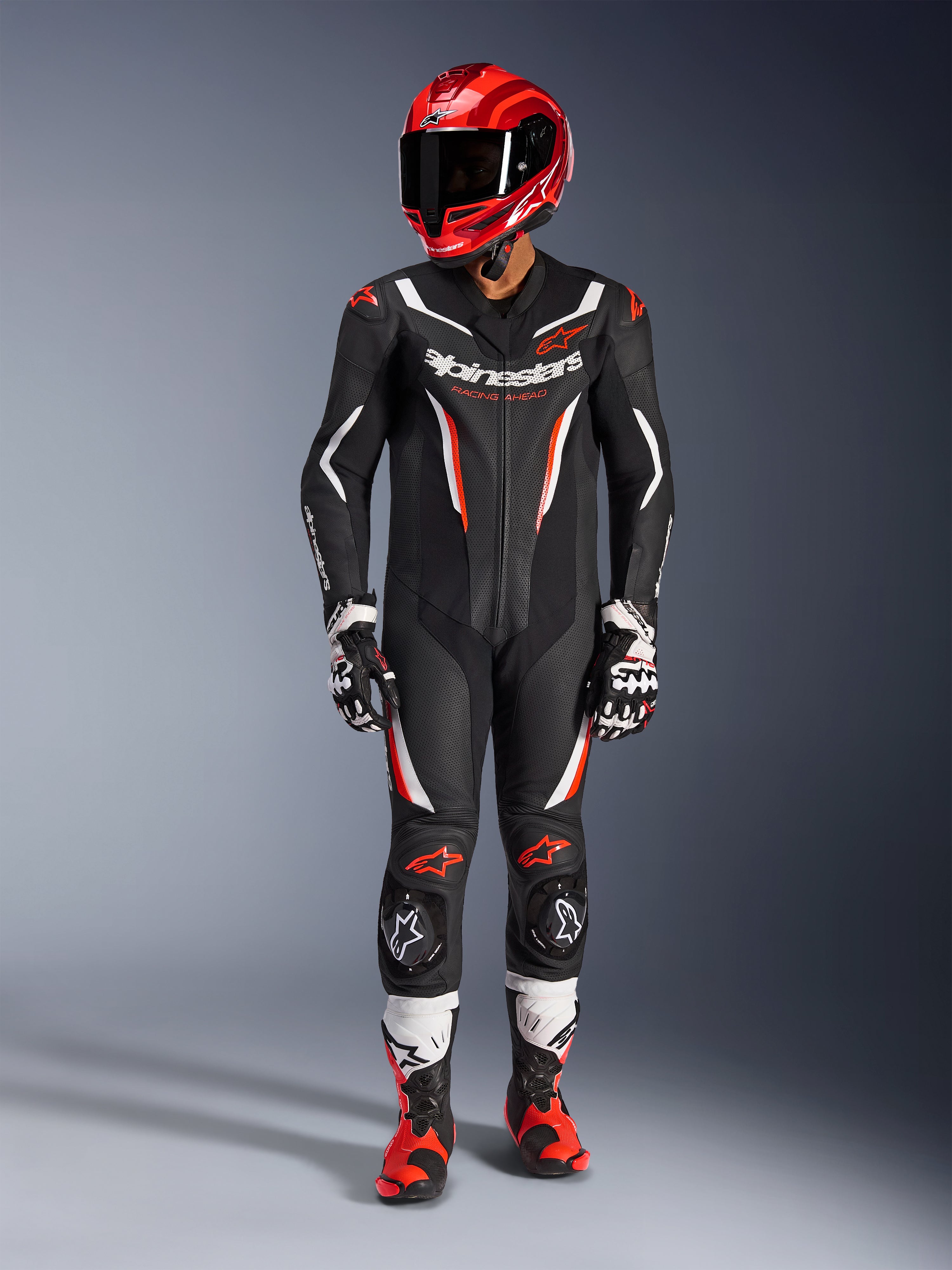 Alpinestars GP Force V2 Leren Pak - 1-delig, Black White Red Fluo, gedragen met een rode racehelm, zwart-witte handschoenen en rood-zwarte laarzen, met geperforeerde leren panelen en Alpinestars-branding, professionele motorrace-uitrusting