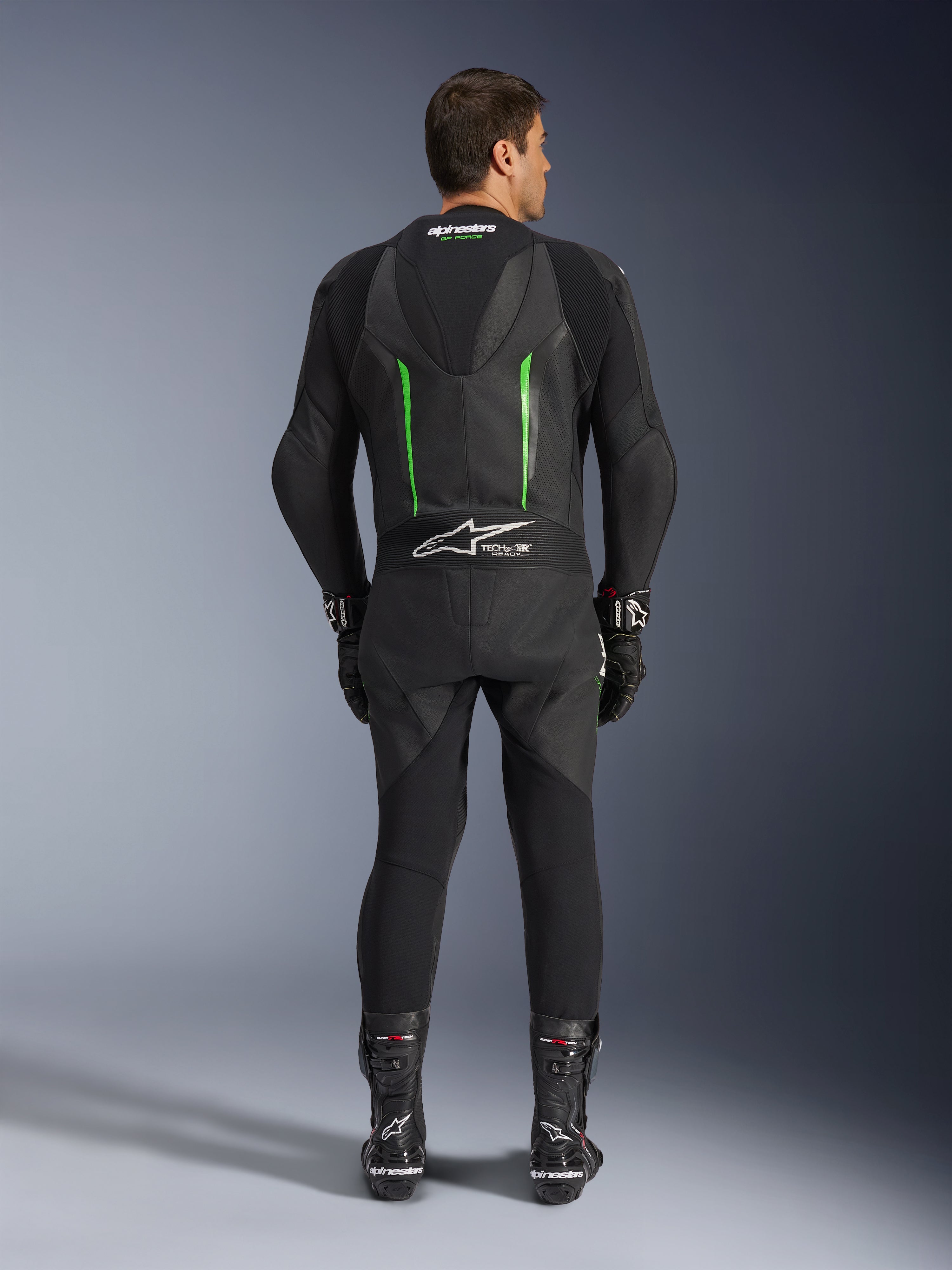 Achteraanzicht van het Alpinestars GP Force V2 1-delig leren pak in zwart en groen, voorzien van een aerodynamische speed hump, geperforeerde panelen en Tech-Air Ready-branding op de onderrug, gedragen met zwarte racehandschoenen en laarzen.
