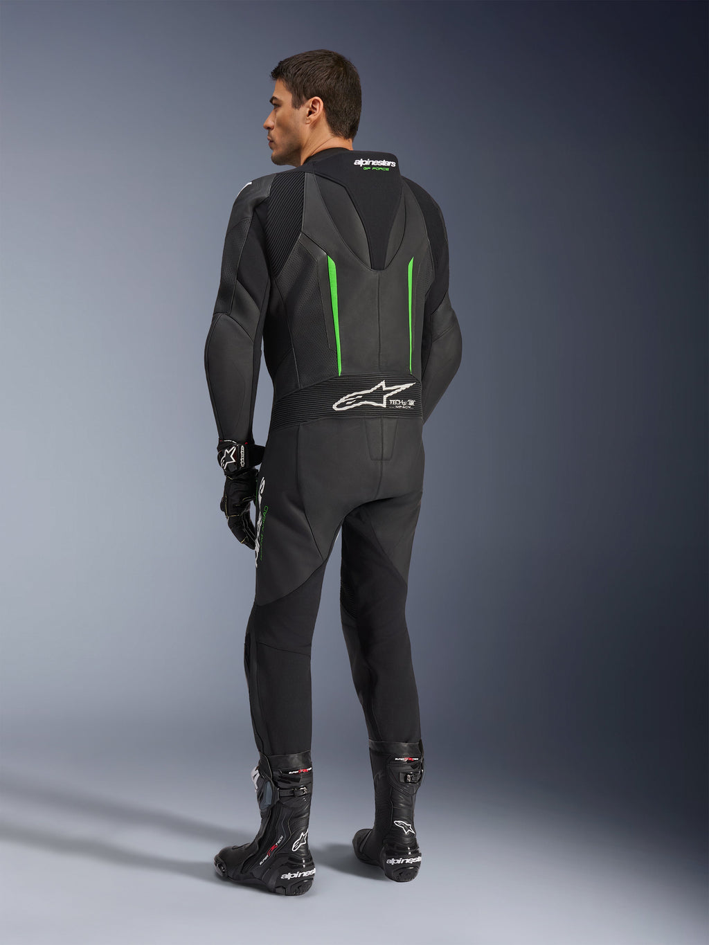 Alpinestars GP Force V2 Leren Pak, 1-delig motorpak, zwart met groene accenten, achteraanzicht met de aerodynamische speed hump en wit logo op het accordeon stretchpaneel, gecombineerd met zwarte racelaarzen en handschoenen.