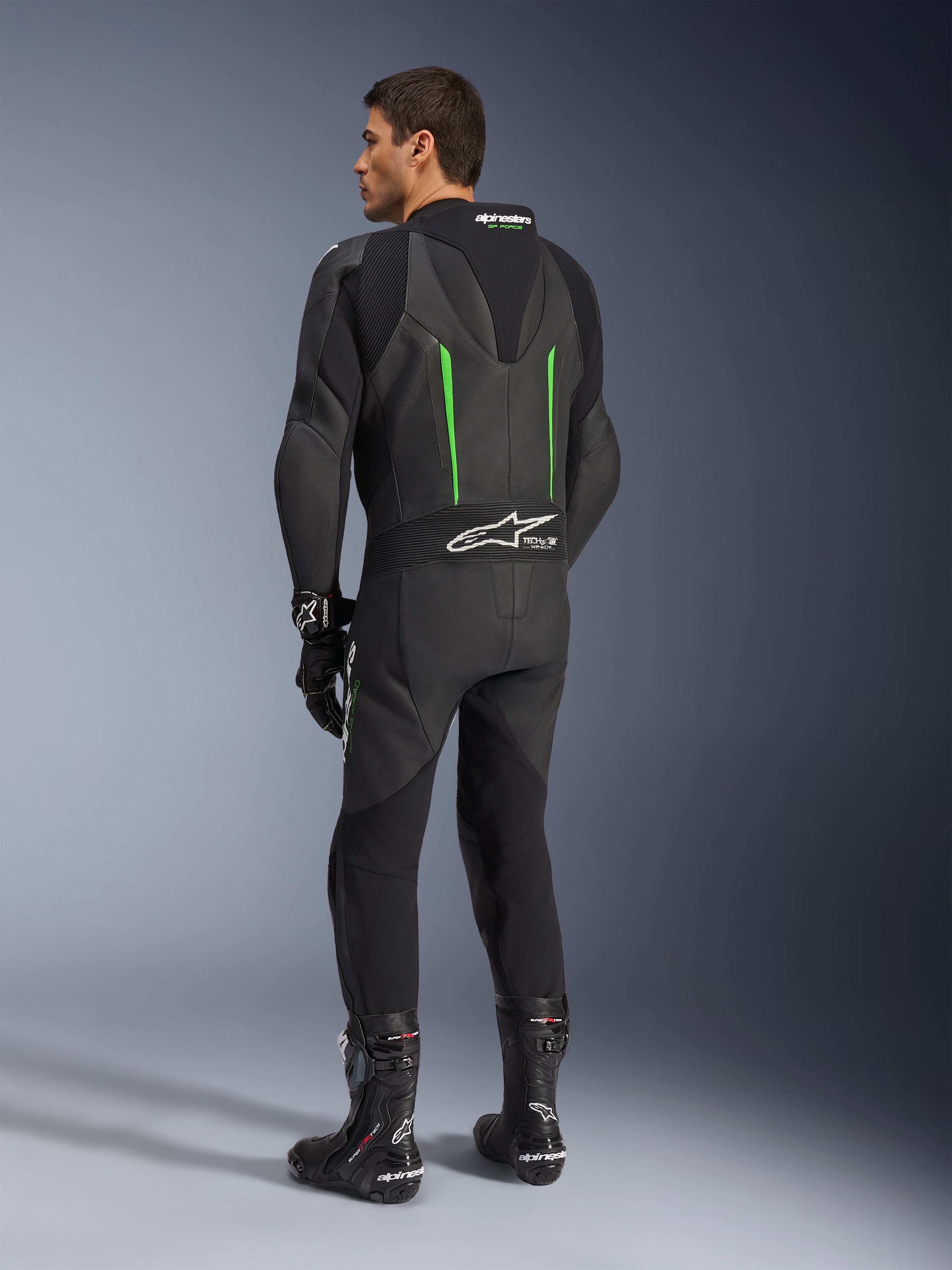 Alpinestars GP Force V2 Leren Pak, 1-delig motorpak, zwart met groene accenten, achteraanzicht met de aerodynamische speed hump en wit logo op het accordeon stretchpaneel, gecombineerd met zwarte racelaarzen en handschoenen.