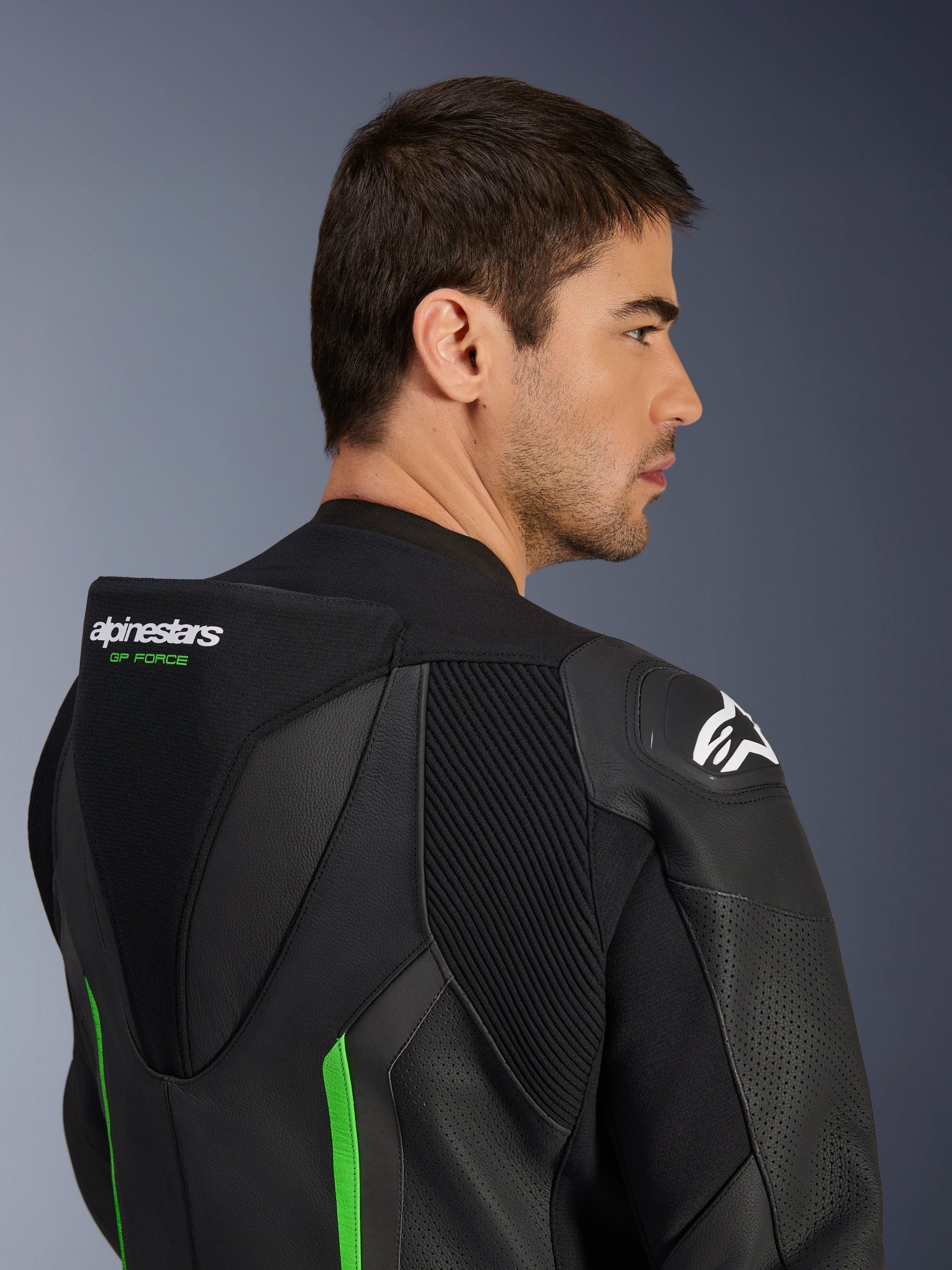 Close-up van de bovenrug en schouder van een mannelijk model in het Alpinestars GP Force V2 Leren Pak in zwart en groen, voorzien van de aerodynamische speed hump met groene 'GP FORCE' branding, een wit Astars-logo op de schouder en accordeon stretchpanelen.