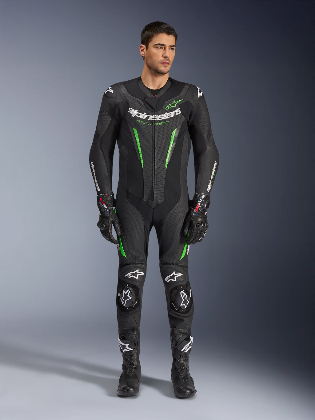 Alpinestars GP Force V2 Leren Pak - 1-delig, eendelig motorracepak, zwart en groen, vooraanzicht van een model dat het pak draagt met witte logo's op de borst en neongroene accenten, voorzien van geperforeerd leer en externe schoudersliders, gecombineerd met zwarte racehandschoenen en laarzen