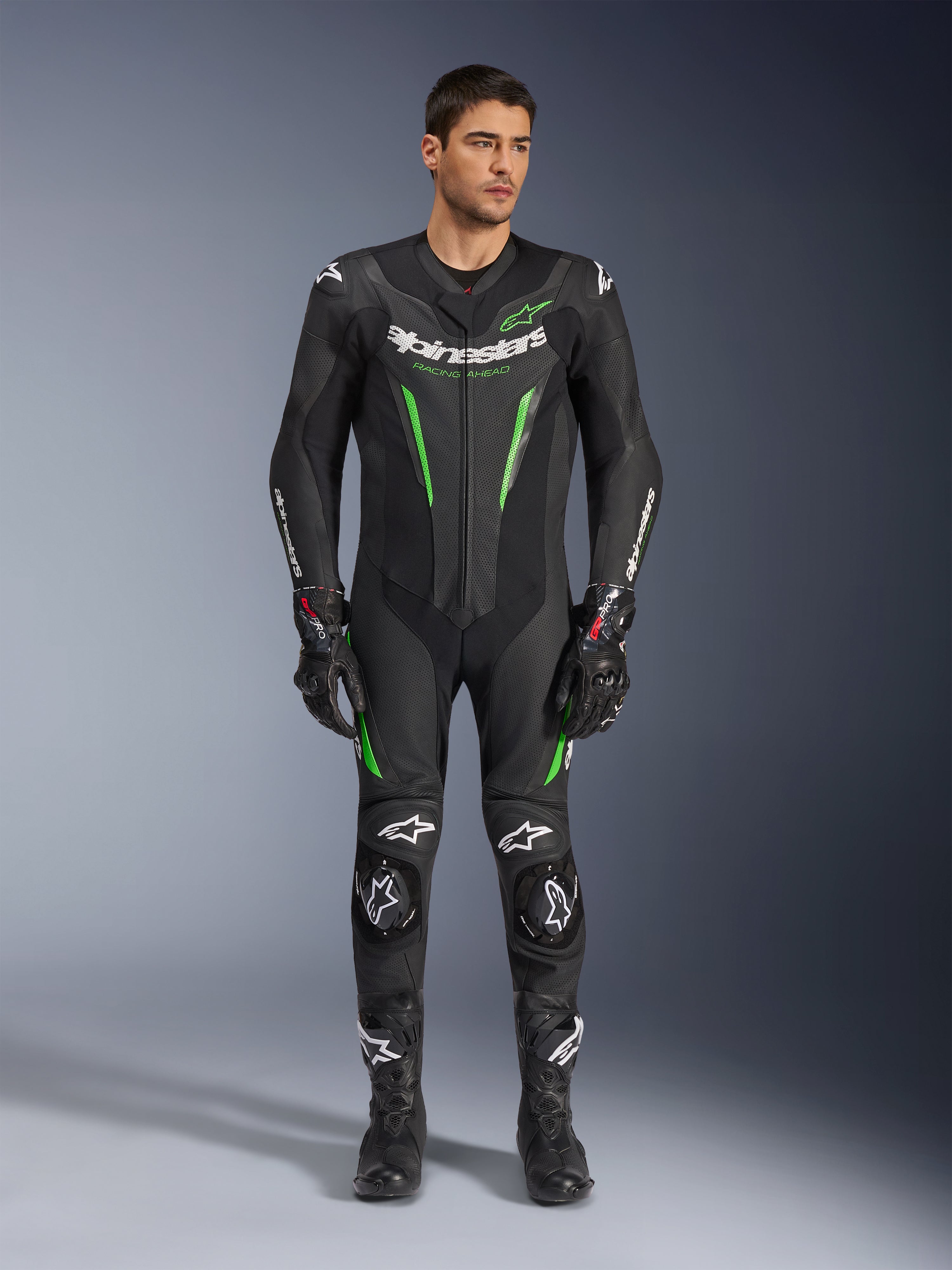 Alpinestars GP Force V2 Leren Pak - 1-delig, eendelig motorracepak, zwart en groen, vooraanzicht van een model dat het pak draagt met witte logo's op de borst en neongroene accenten, voorzien van geperforeerd leer en externe schoudersliders, gecombineerd met zwarte racehandschoenen en laarzen