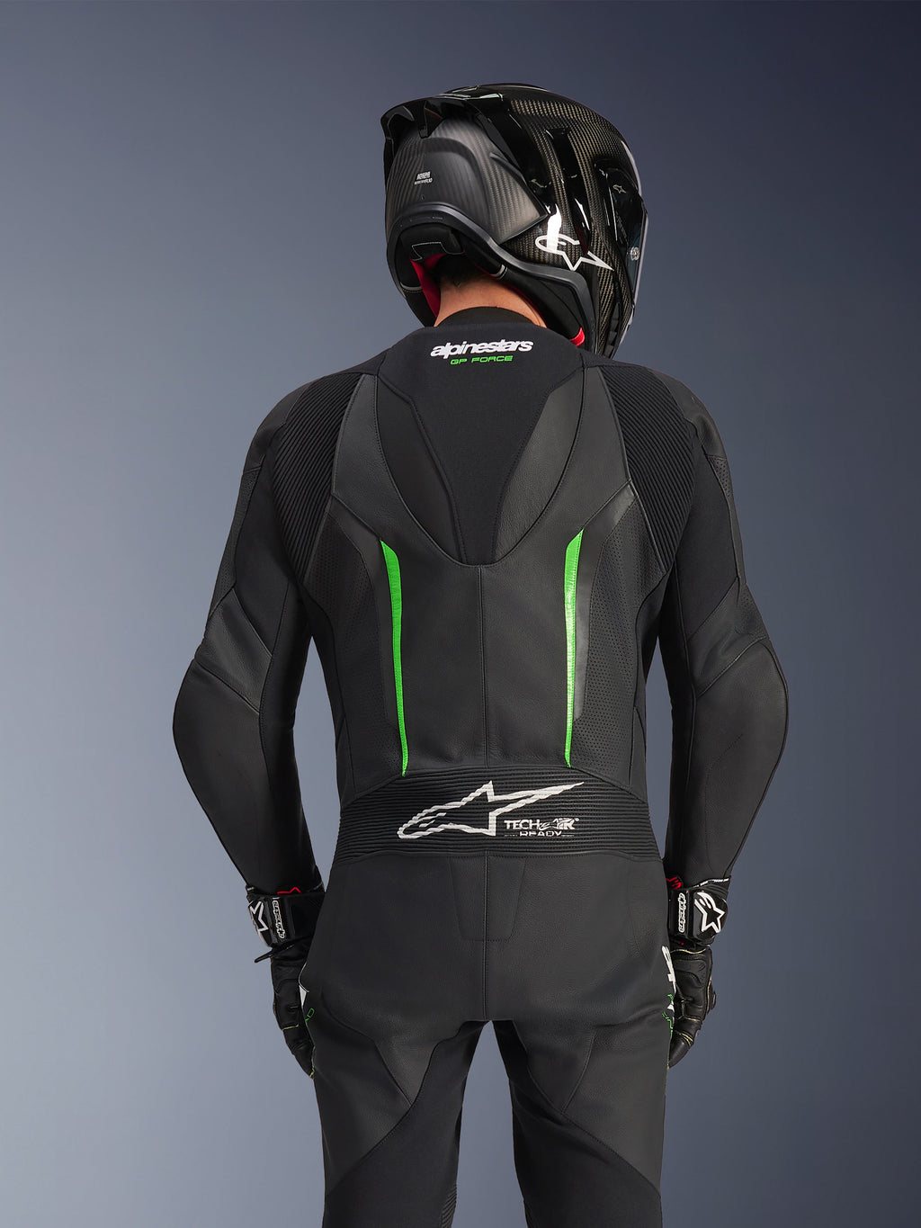 Achteraanzicht van een rijder die het Alpinestars GP Force V2 Leren Pak in Black Green draagt, voorzien van een aerodynamische speed hump, Tech-Air Ready-branding op het accordeonpaneel op de onderrug en een geperforeerde lederen constructie, gecombineerd met een zwarte carbon racehelm.