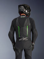 Achteraanzicht van een rijder die het Alpinestars GP Force V2 Leren Pak in Black Green draagt, voorzien van een aerodynamische speed hump, Tech-Air Ready-branding op het accordeonpaneel op de onderrug en een geperforeerde lederen constructie, gecombineerd met een zwarte carbon racehelm.