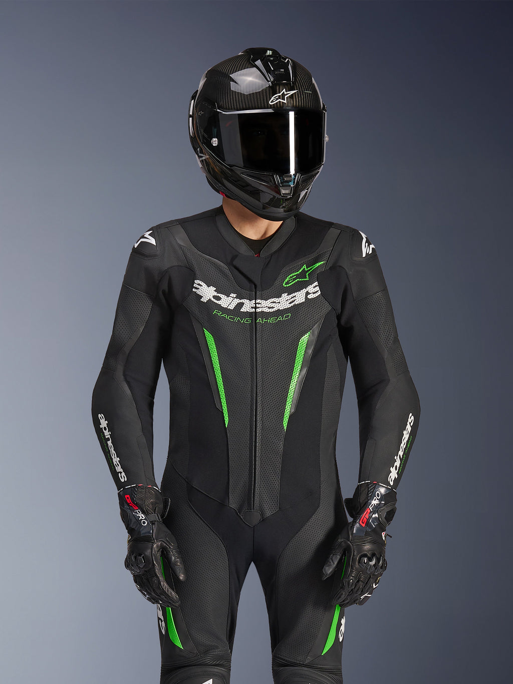 Alpinestars GP Force V2 Leren Pak - 1-delig, zwart en groen, motorracepak gedragen met een carbon fiber helm en GP Pro-handschoenen, voorzien van limoengroene accenten en Alpinestars 'Racing Ahead'-branding, ontworpen voor circuitprestaties.