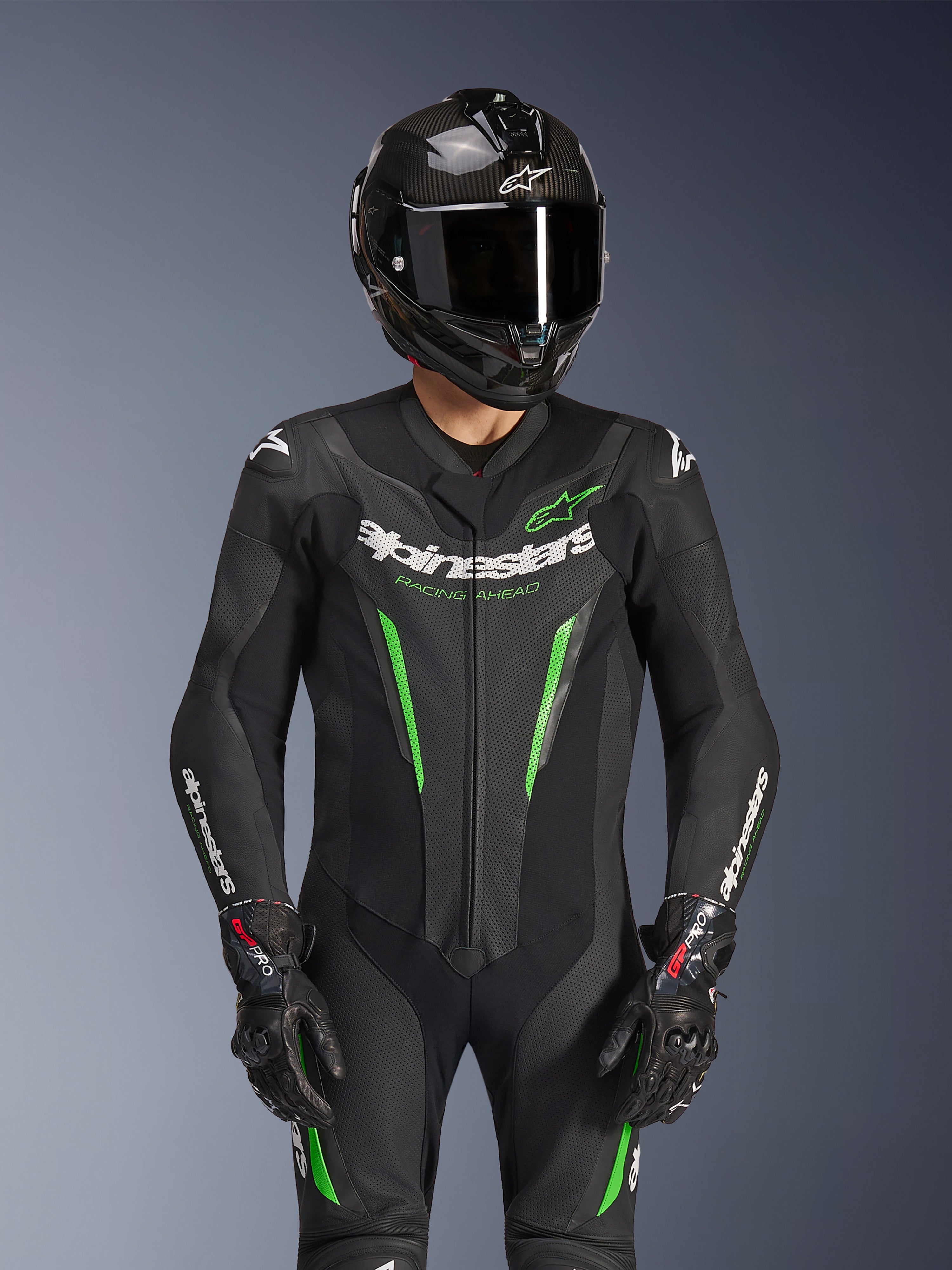 Alpinestars GP Force V2 Leren Pak - 1-delig, zwart en groen, motorracepak gedragen met een carbon fiber helm en GP Pro-handschoenen, voorzien van limoengroene accenten en Alpinestars 'Racing Ahead'-branding, ontworpen voor circuitprestaties.