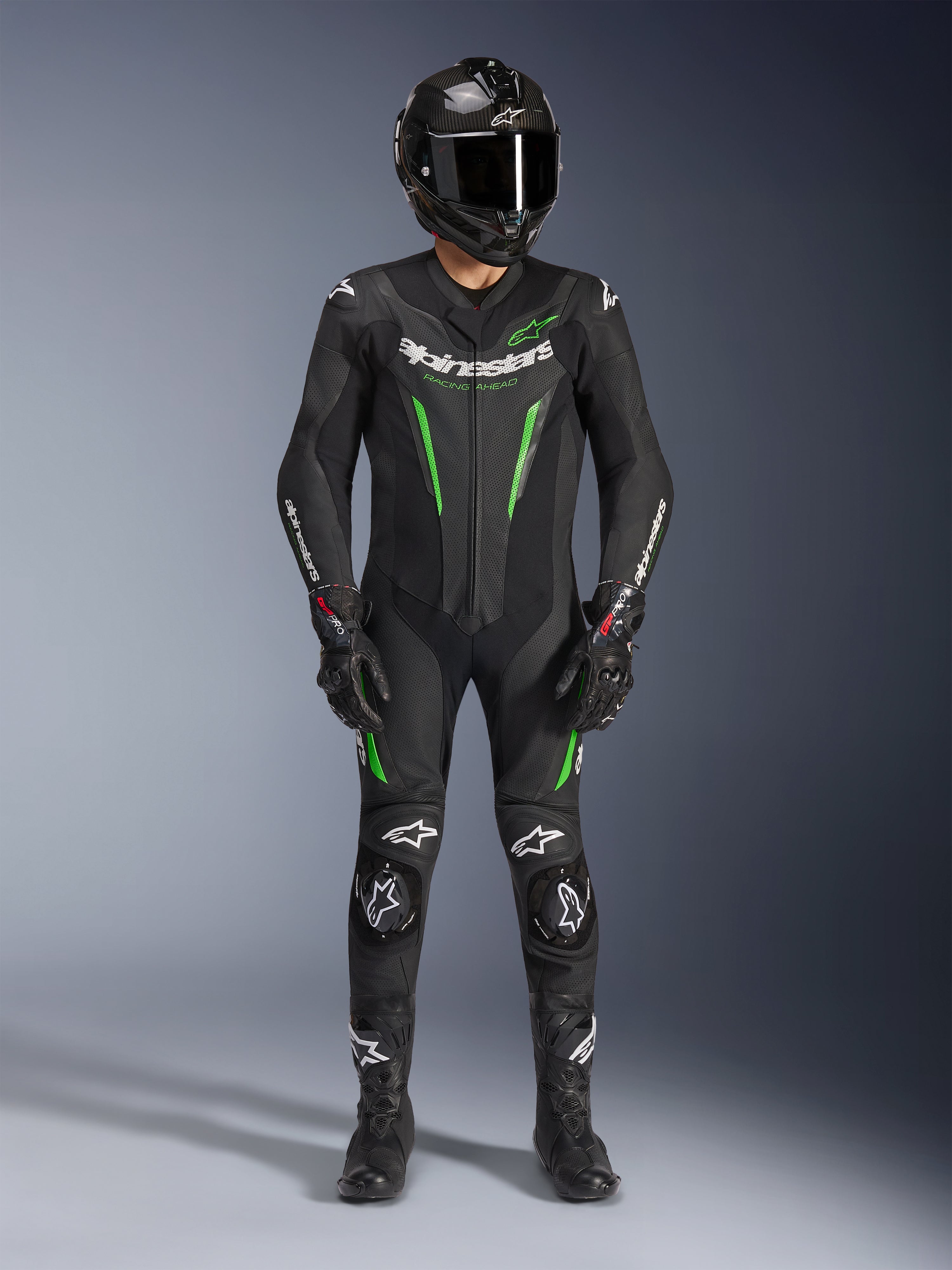 Motorrijder draagt Alpinestars GP Force V2 Leren Pak - 1-delig in zwart en groen, gecombineerd met een carbon fiber helm en racehandschoenen, voorzien van geperforeerde leren panelen en witte Alpinestars-logo's, professionele uitrusting voor circuit en sportief rijden