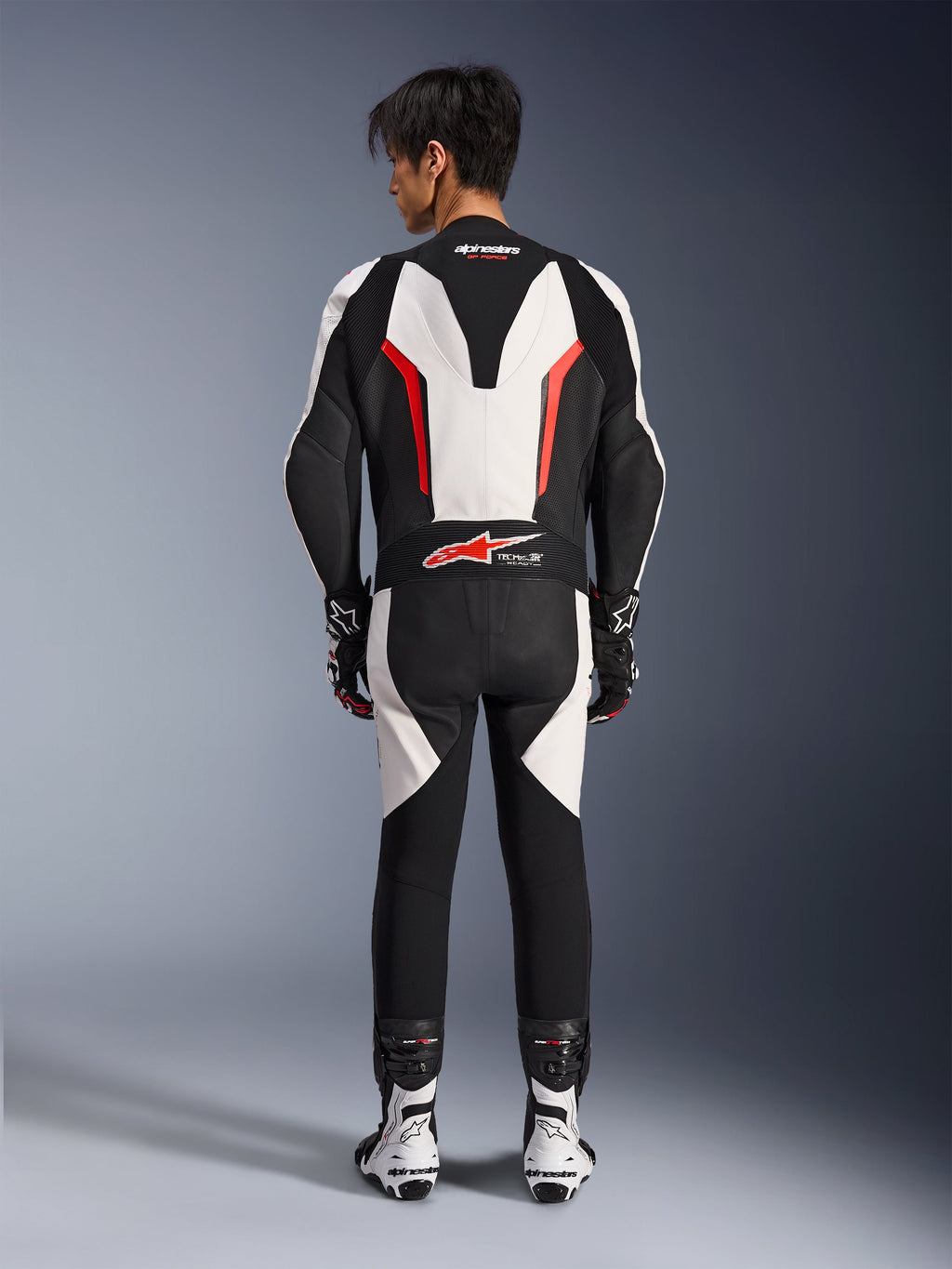 Alpinestars GP Force V2 eendelig leren pak, motorracepak, wit, zwart en fluo rood, achteraanzicht van model dat het pak draagt met racelaarzen en handschoenen, voorzien van een aerodynamische speedhump, accordeon stretchpanelen en Tech-Air Ready branding