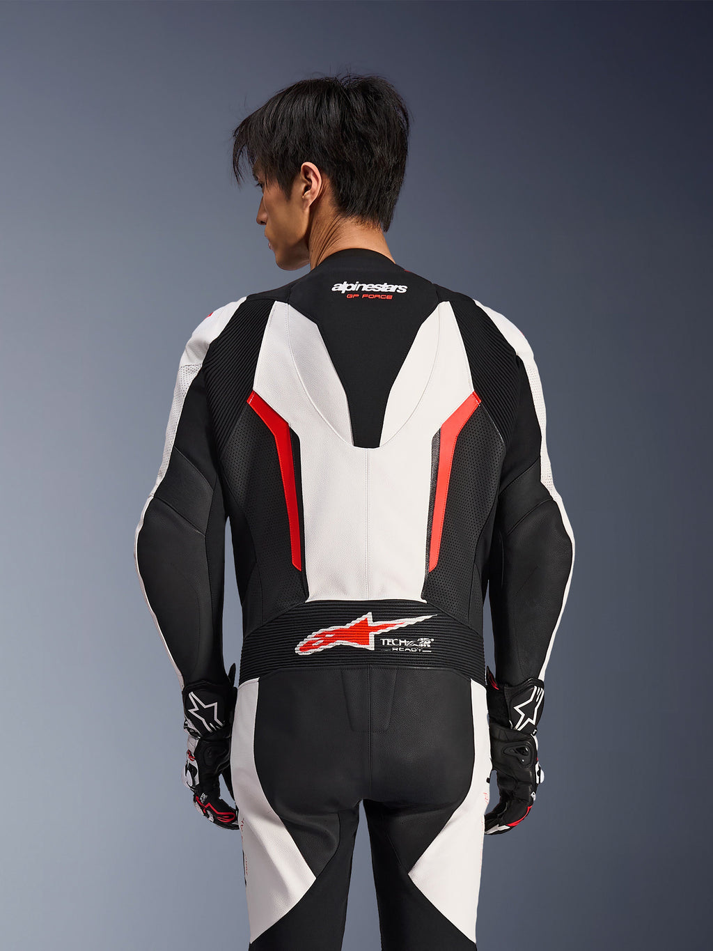 Alpinestars GP Force V2 Leren Pak 1-delig, wit, zwart en fluo rood, achteraanzicht op mannelijk model, met aerodynamische speed hump, geperforeerde leren panelen en Tech-Air Ready-logo op de onderrug, professionele race-uitrusting met zwart-witte handschoenen