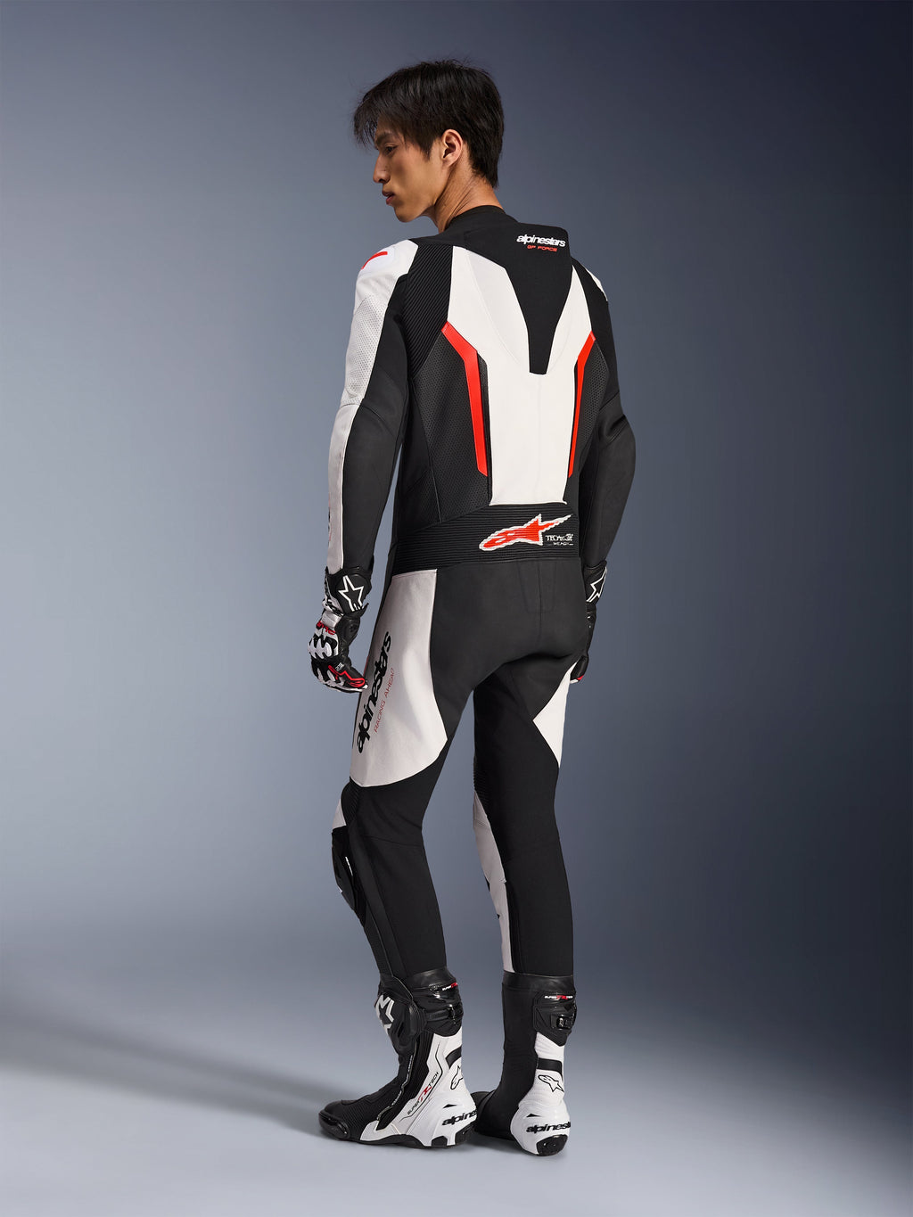 Alpinestars GP Force V2 Leren Pak - 1-delig in wit, zwart en fluorood, achteraanzicht van een motorrijder die de aerodynamische speed hump en geperforeerde leren panelen toont, gecombineerd met bijpassende racelaarzen en handschoenen, ontworpen voor het circuit.