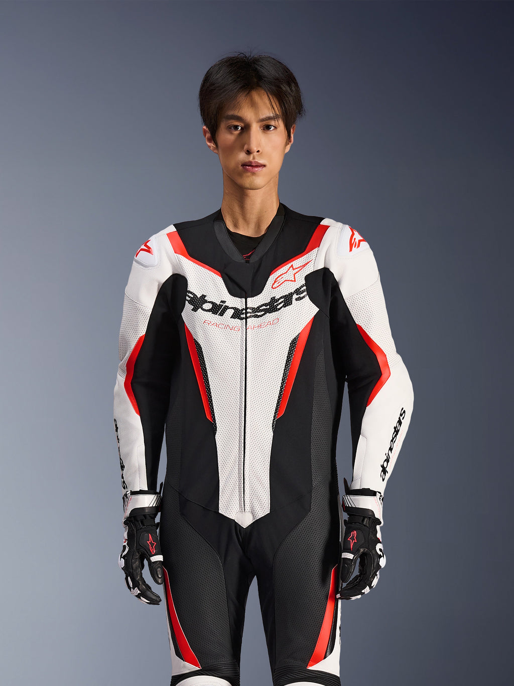 Alpinestars GP Force V2 Leren Pak - 1-delig, Wit Zwart en Rood Fluo, gedragen door een mannelijk model, voorzien van geperforeerde leren panelen en opvallende Alpinestars-branding op de borst, gecombineerd met zwart-witte racehandschoenen, ontworpen voor circuit- en prestatiegerichte ritten