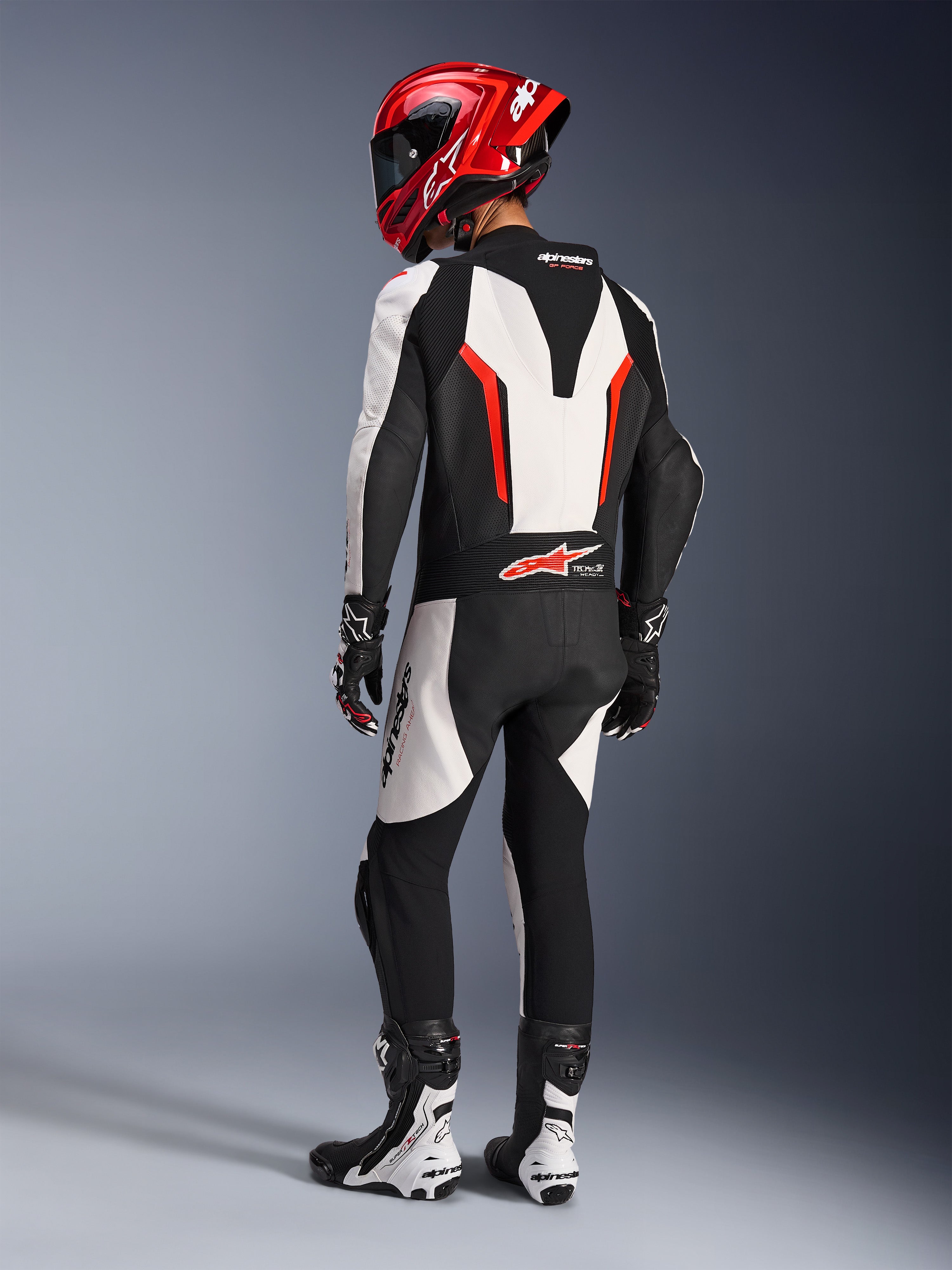 Alpinestars GP Force V2 Leren Pak - 1-delig, wit zwart rood fluo, achteraanzicht van rijder in volledige race-uitrusting, inclusief rode helm, zwart-witte laarzen en handschoenen, waarbij de aerodynamische speedhump en ergonomische stretchpanelen worden benadrukt.