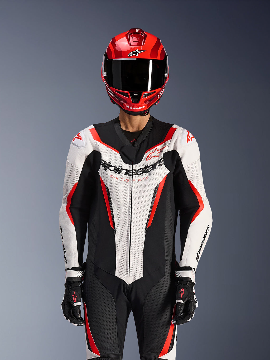 Alpinestars GP Force V2 Leren Pak - 1-delig, wit zwart rood fluo, gedragen met een rode integraalhelm en zwarte racehandschoenen, voorzien van geperforeerde leren panelen en opvallende Alpinestars-branding op de borst, professionele uitrusting voor motorraces